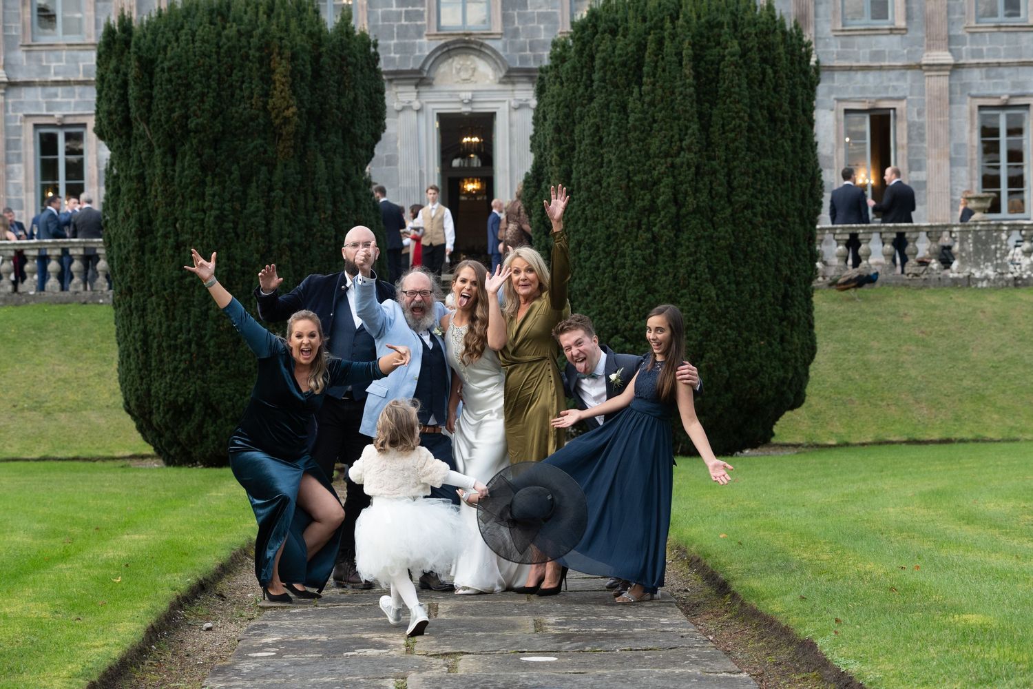 Wedding party posing joyfully outside a grand estate, with guests in formal attire celebrating on a stone pathway.