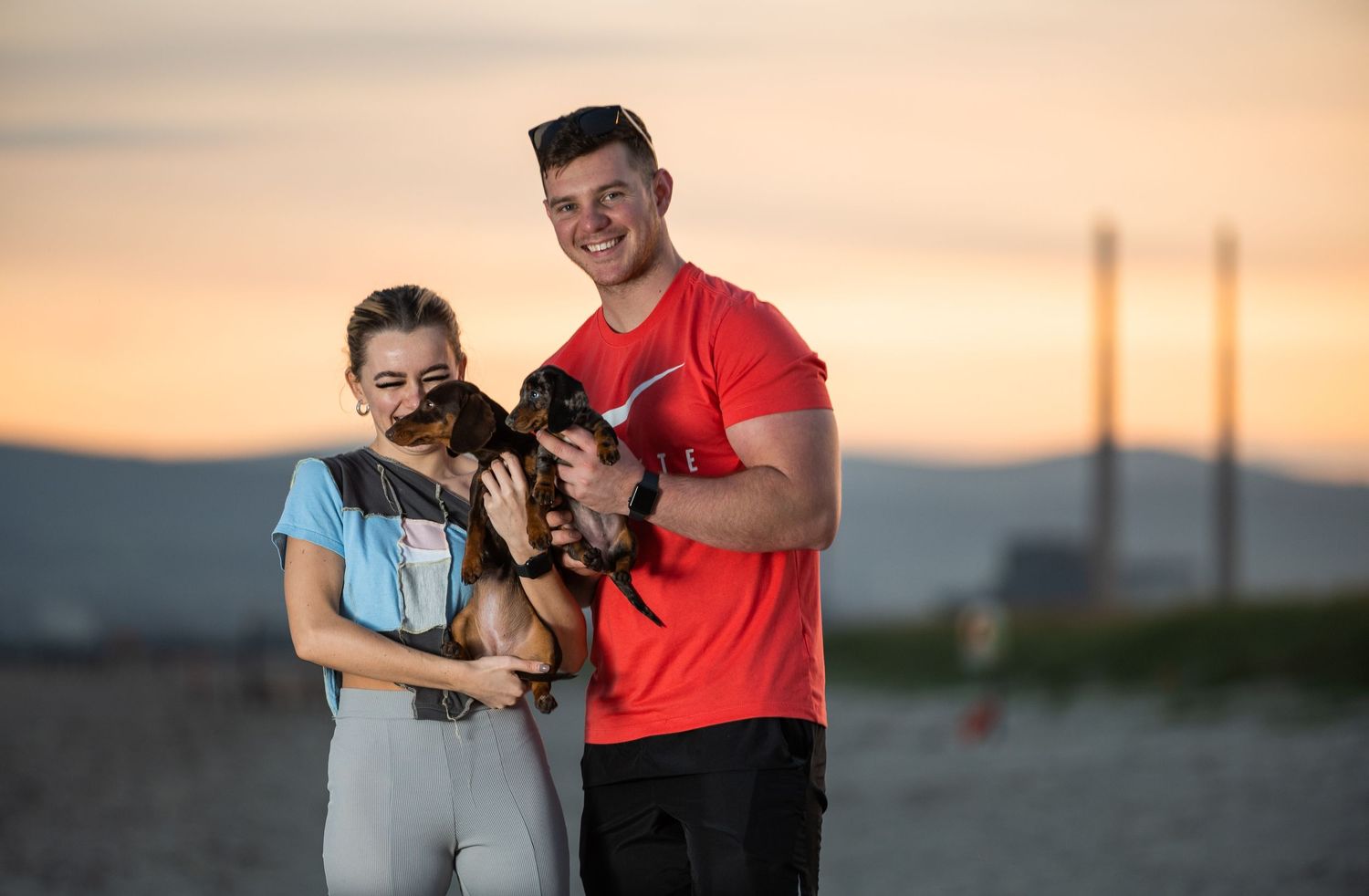 Couple holding small dogs on a beach at sunset, man in red shirt and woman in blue top with gray leggings.