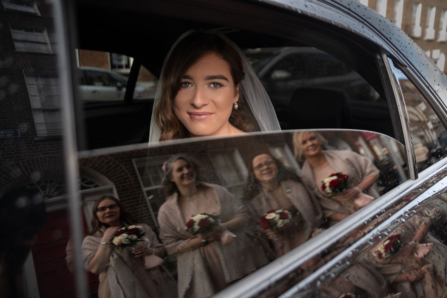 Bride in white veil sits in vintage car, with bridesmaids holding bouquets reflected in the window.