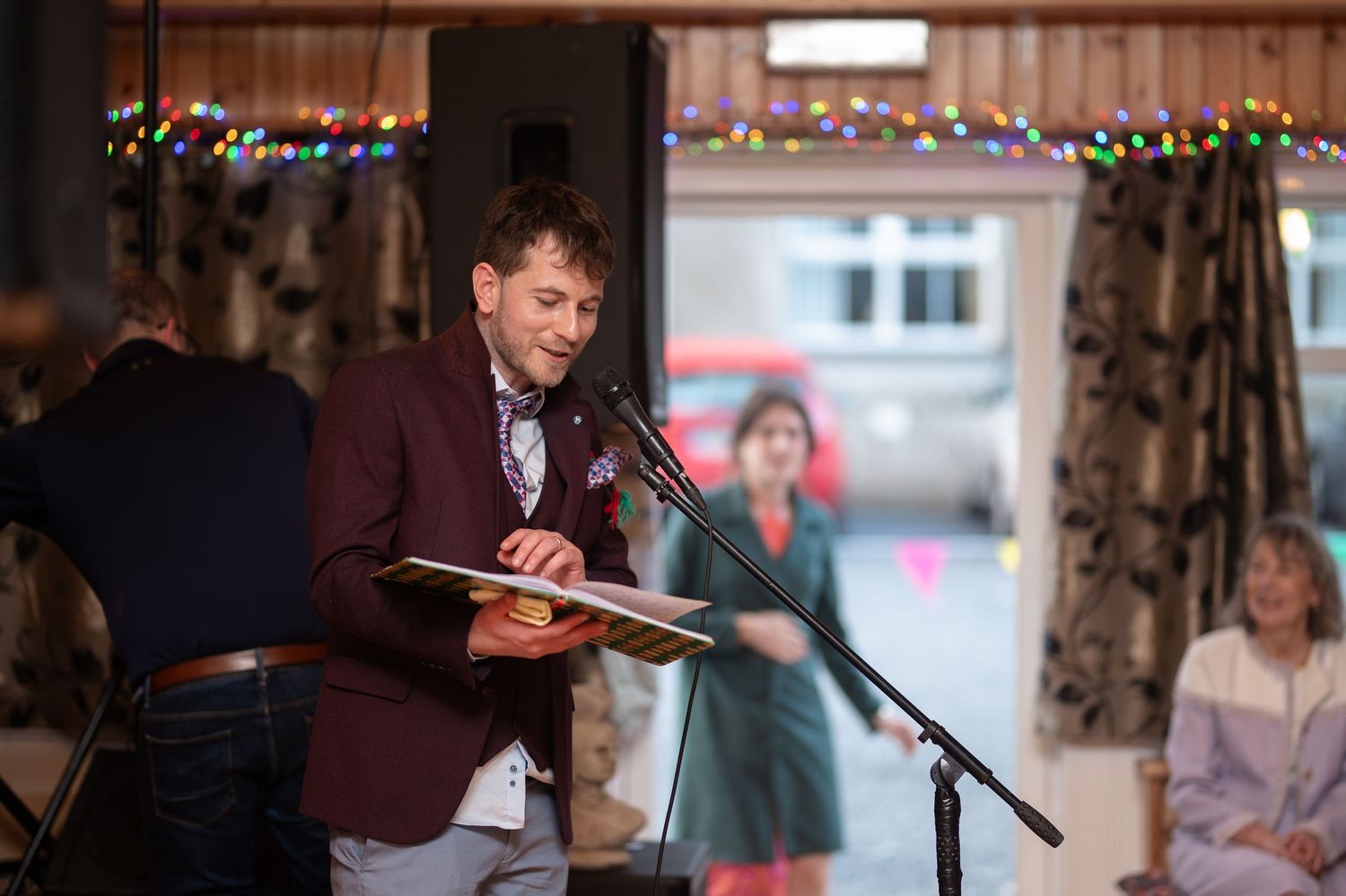 Man in burgundy blazer reads speech at microphone during indoor wedding reception with festive lights.
