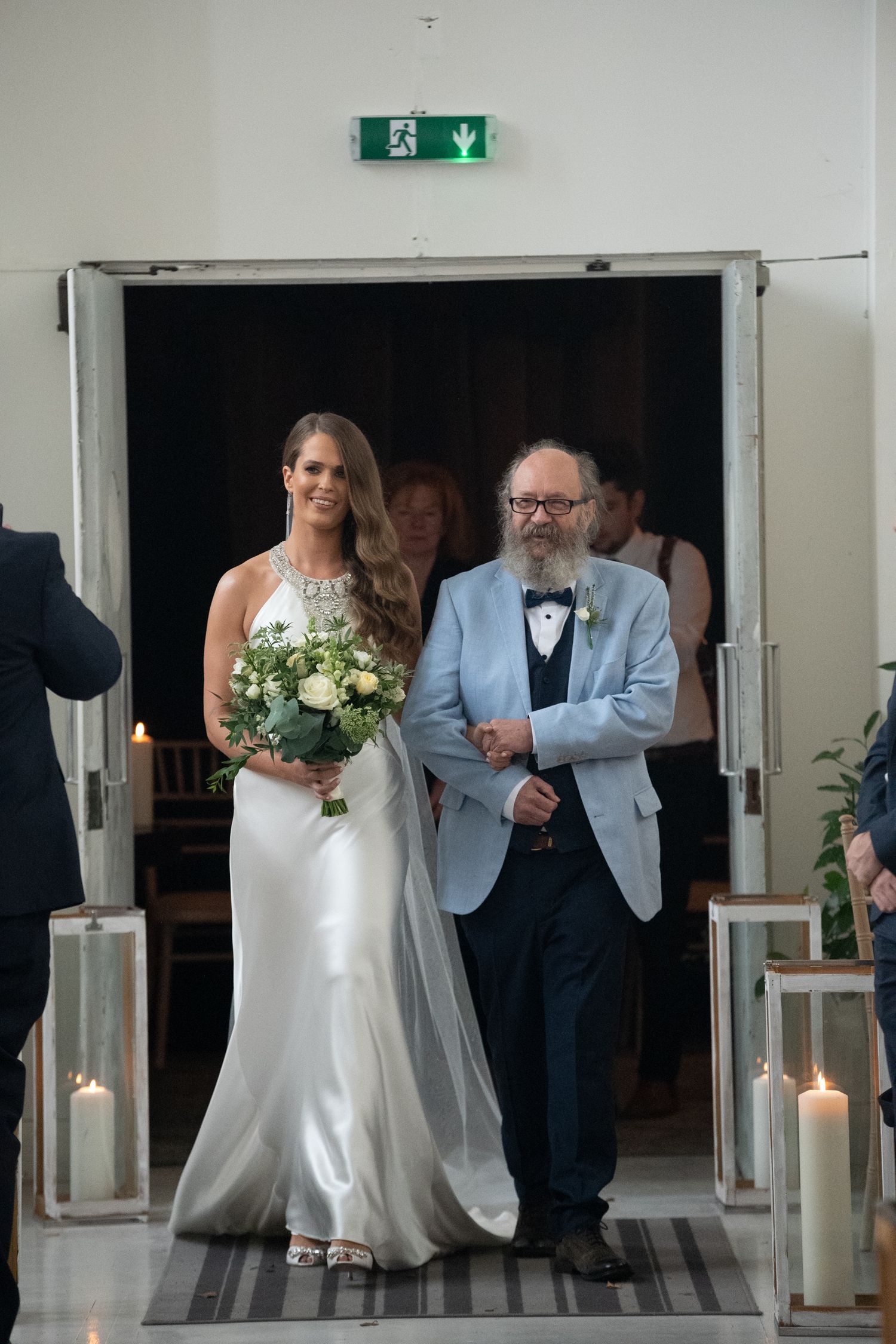 Bride in white gown holding bouquet walks down aisle with bearded man in blue suit at wedding ceremony.