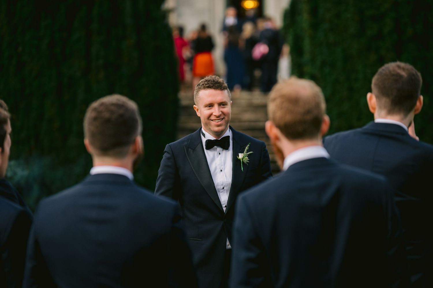Groom in black tuxedo smiling at groomsmen outdoors at wedding ceremony with lush green ivy backdrop.