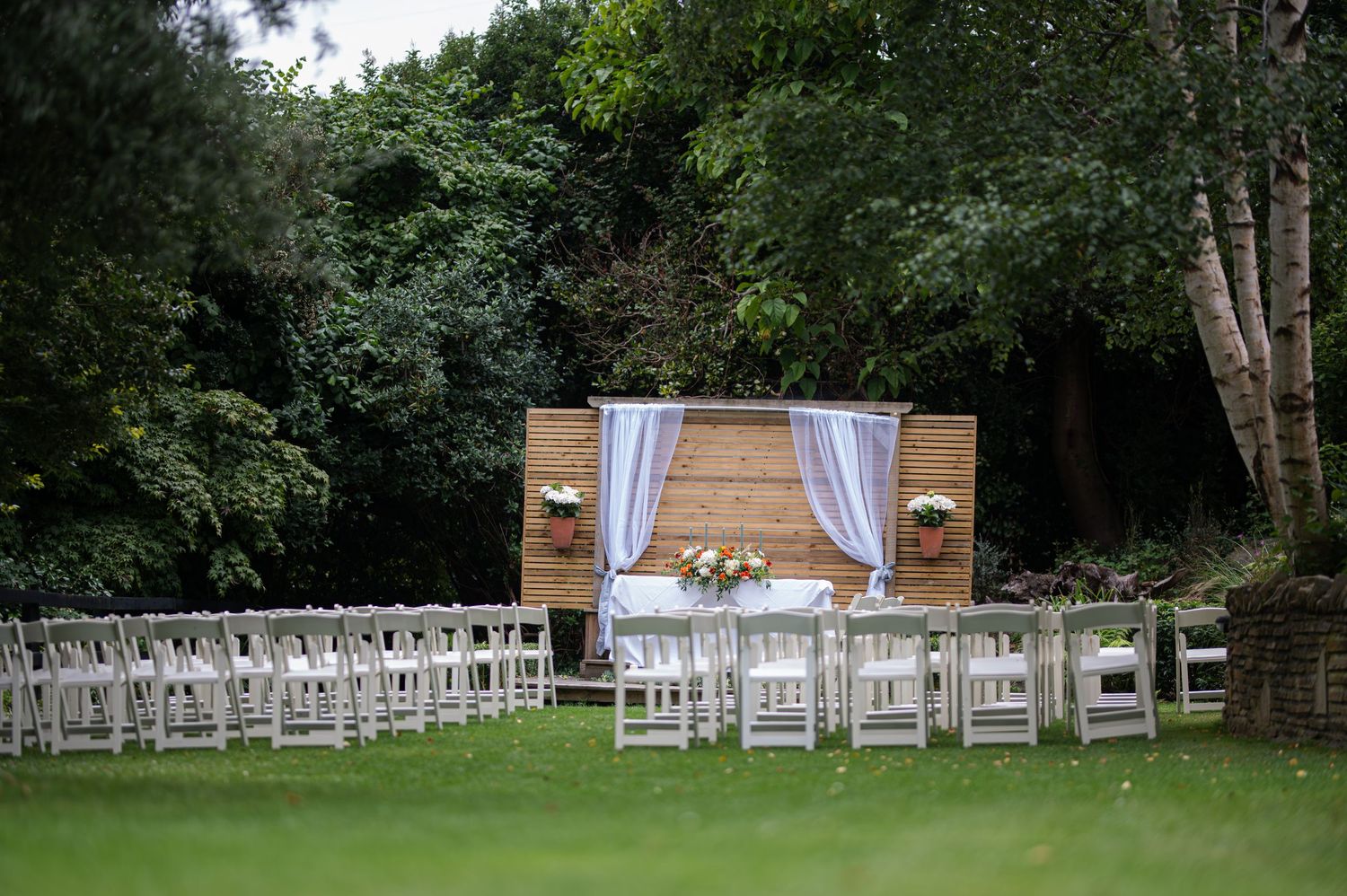 Outdoor wedding ceremony setup with white chairs and wooden altar surrounded by lush green trees.