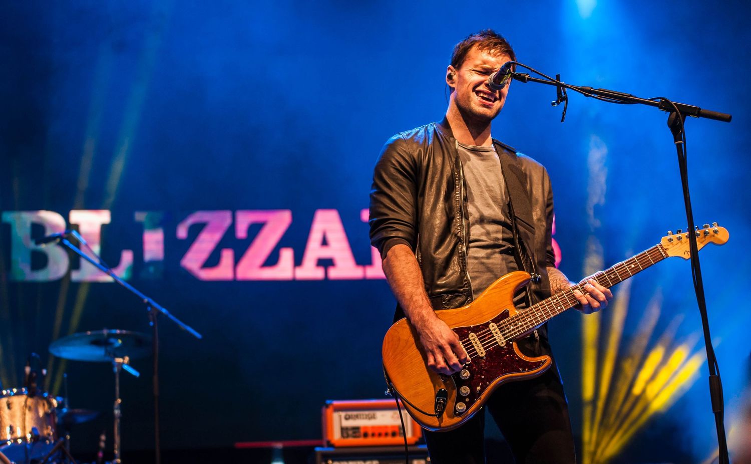 Smiling guitarist performing on stage with Blizzard branding and blue concert lighting.
