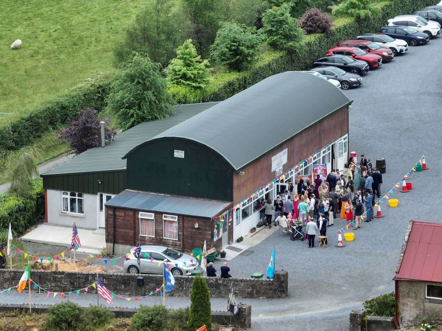 Aerial view of crowd gathered outside rural community hall with parked cars nearby.