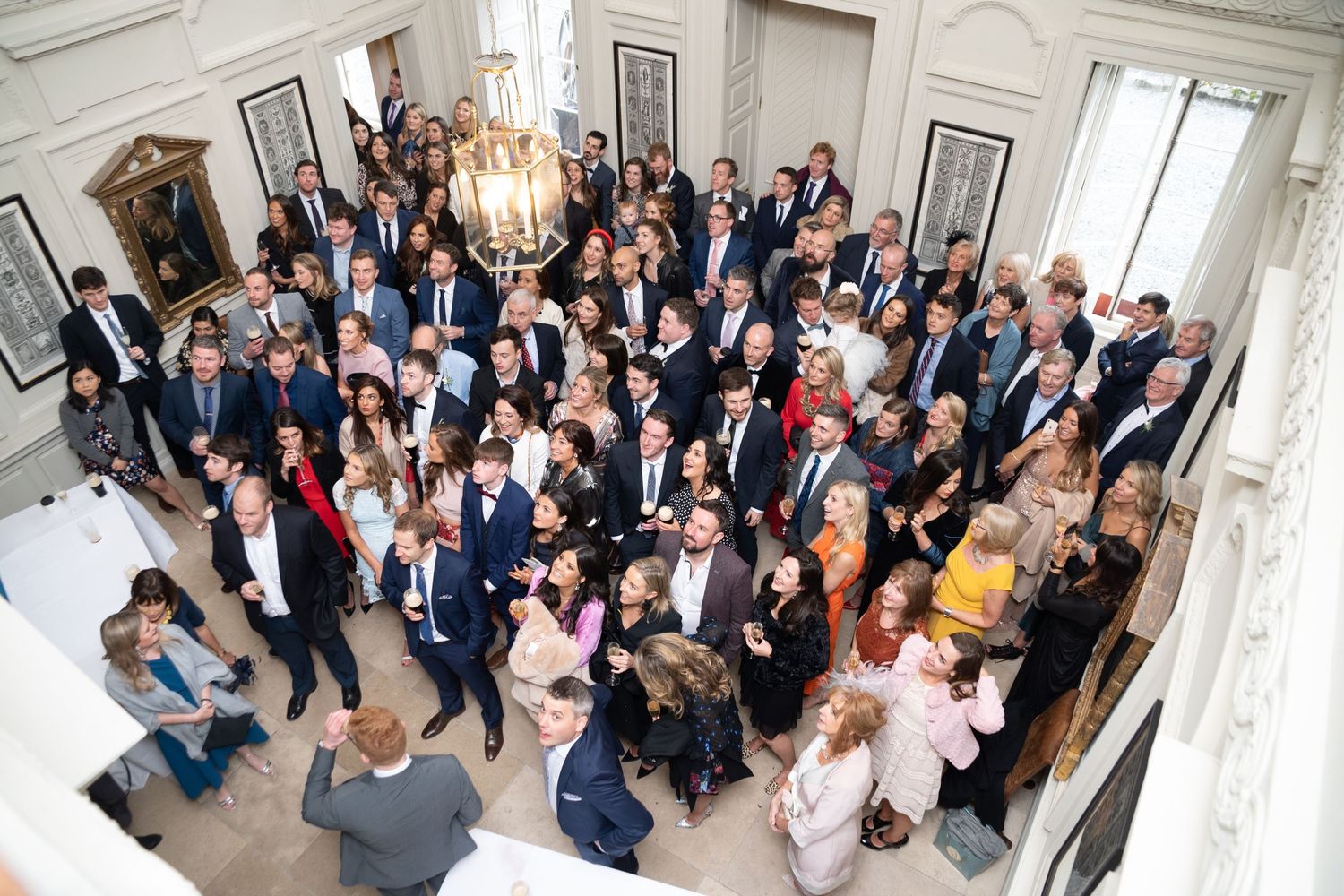 Aerial view of a large elegant wedding reception gathering with guests in formal attire inside a grand venue.