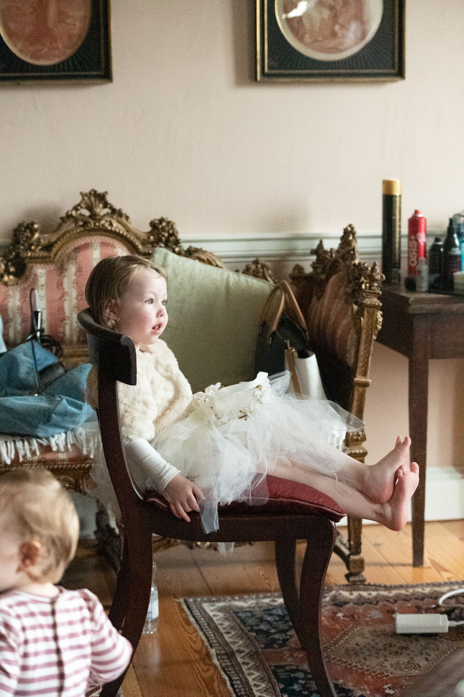 Young girl in white tutu dress sits barefoot on ornate wooden chair in elegant room with framed artwork.