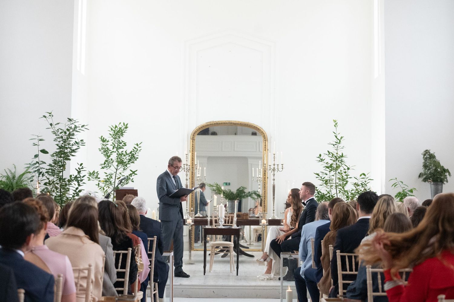 Elegant wedding ceremony in bright white venue with gold mirror, greenery, and guests seated in wooden chairs.
