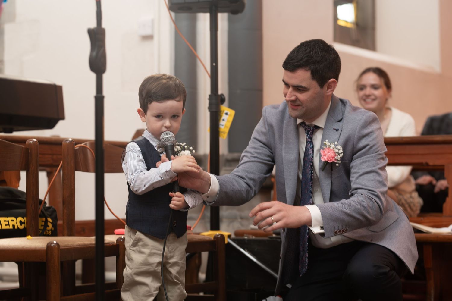 Young boy with pacifier holds microphone assisted by man in gray suit at wedding ceremony.