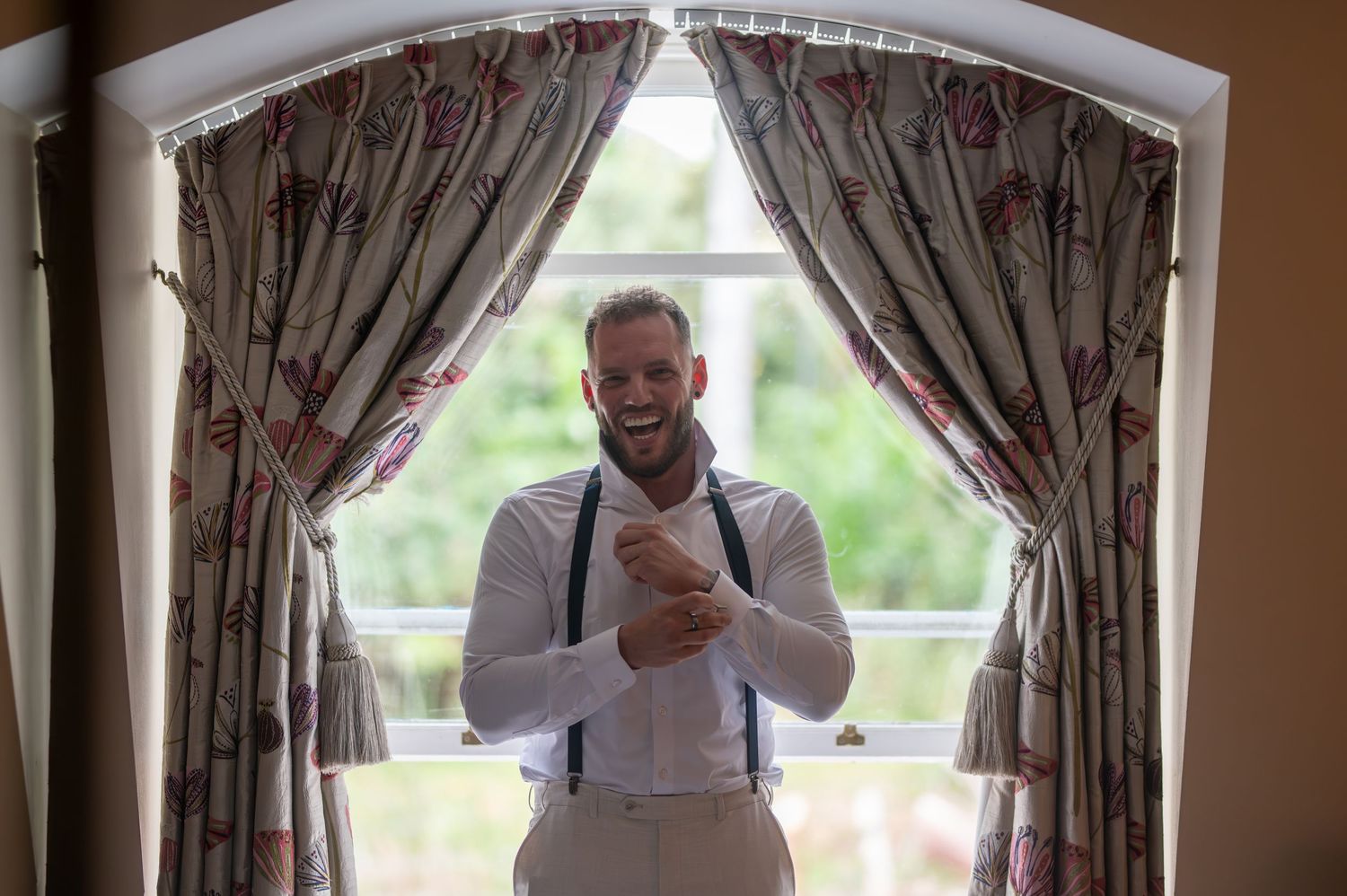 Smiling groom in white shirt and suspenders adjusting cufflinks by a window with floral curtains on his wedding day.