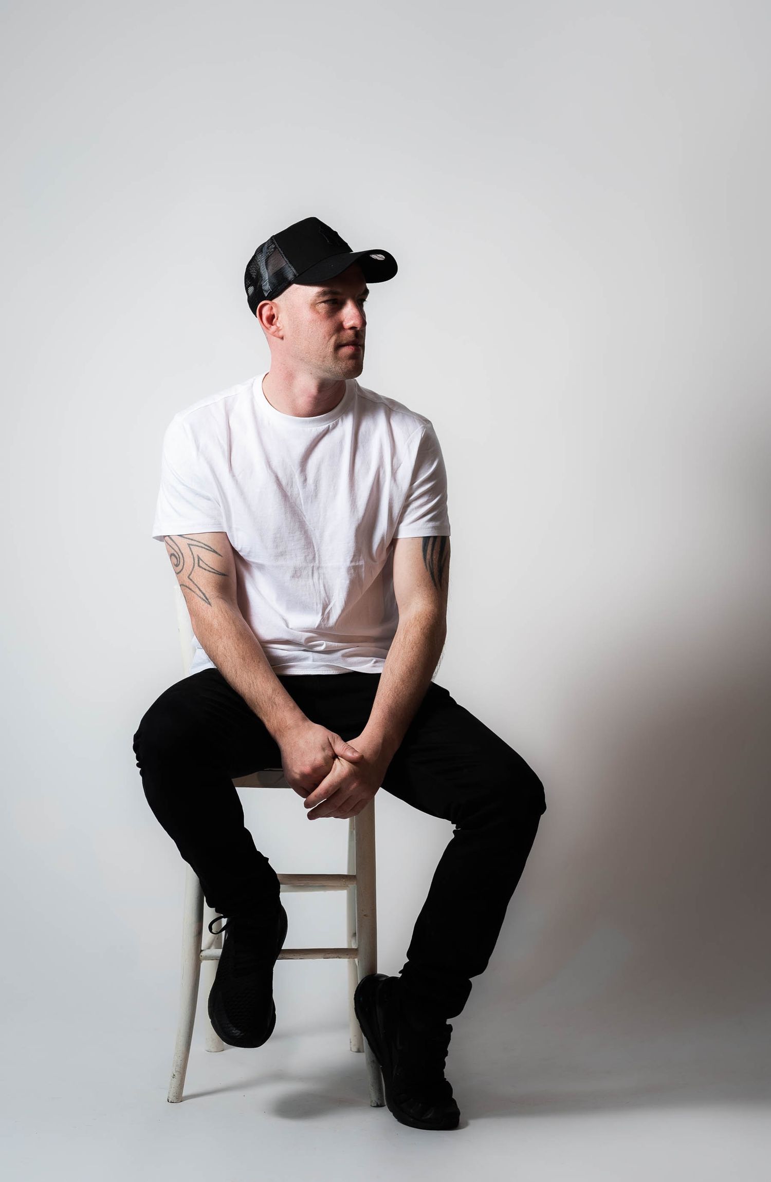 Tattooed man in white t-shirt and black cap sitting on stool against white background.
