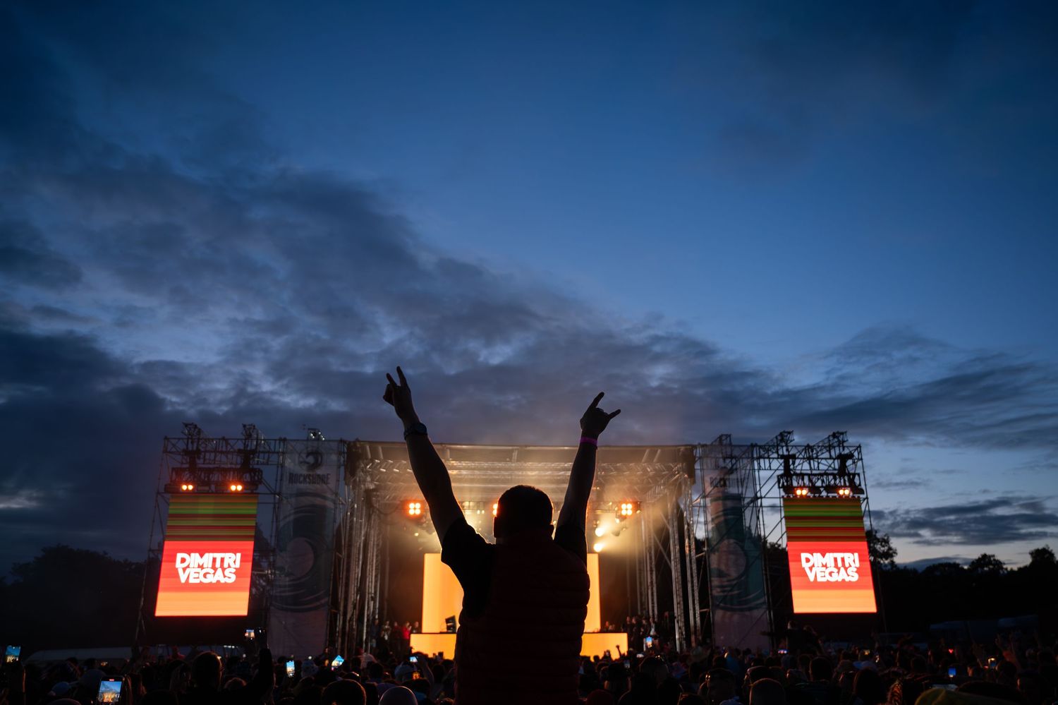 Silhouette of a concert-goer raising hands at an outdoor music festival stage at dusk.