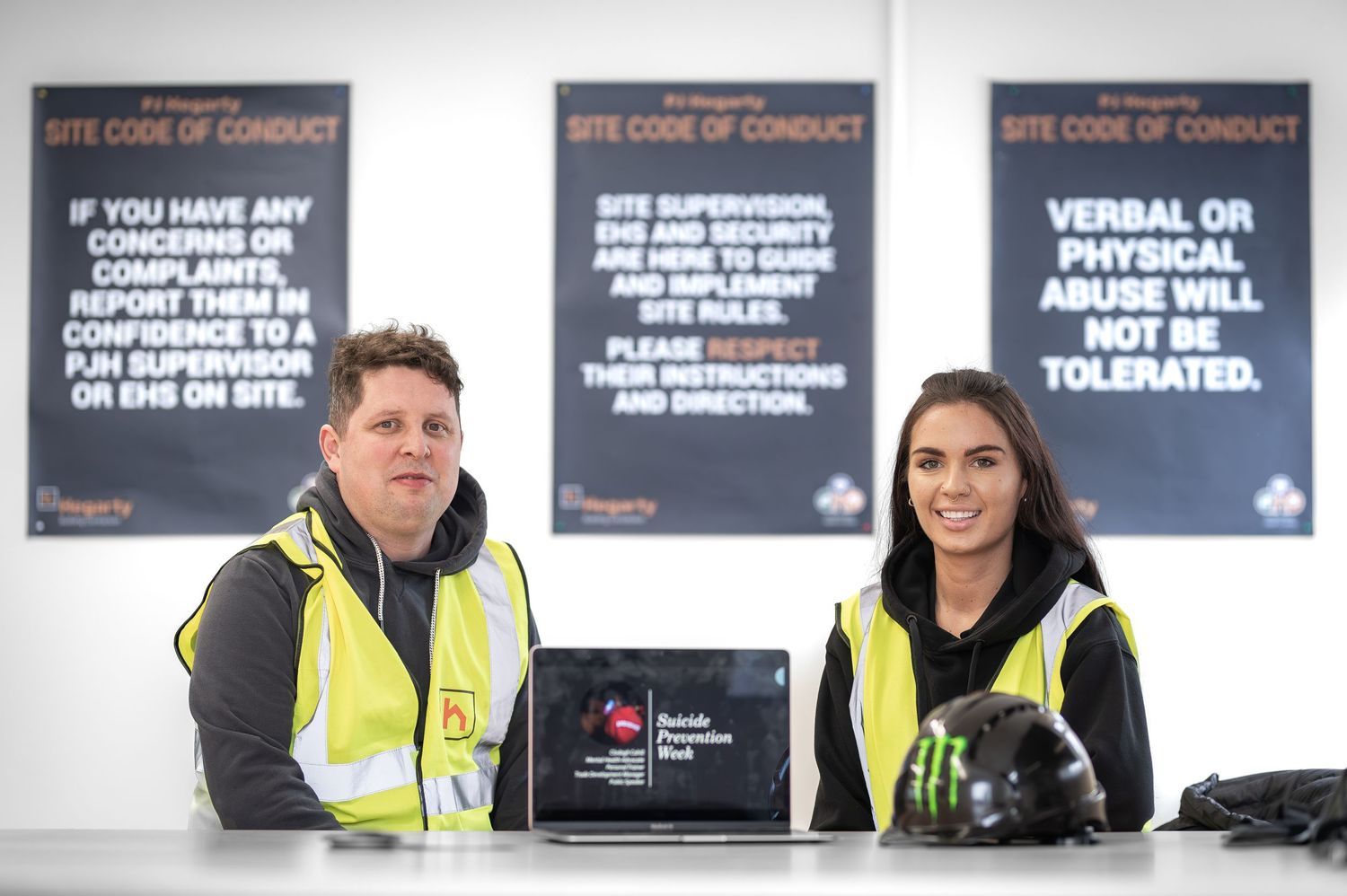 Two workers in hi-vis vests sitting at a registration desk on a construction site.