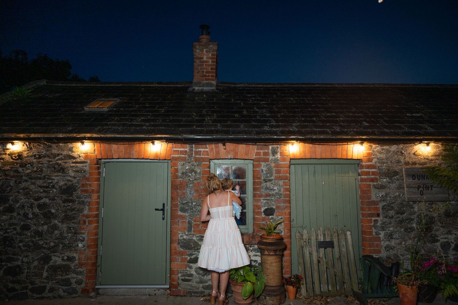 Bride in white dress stands at door of charming stone cottage venue illuminated at night.