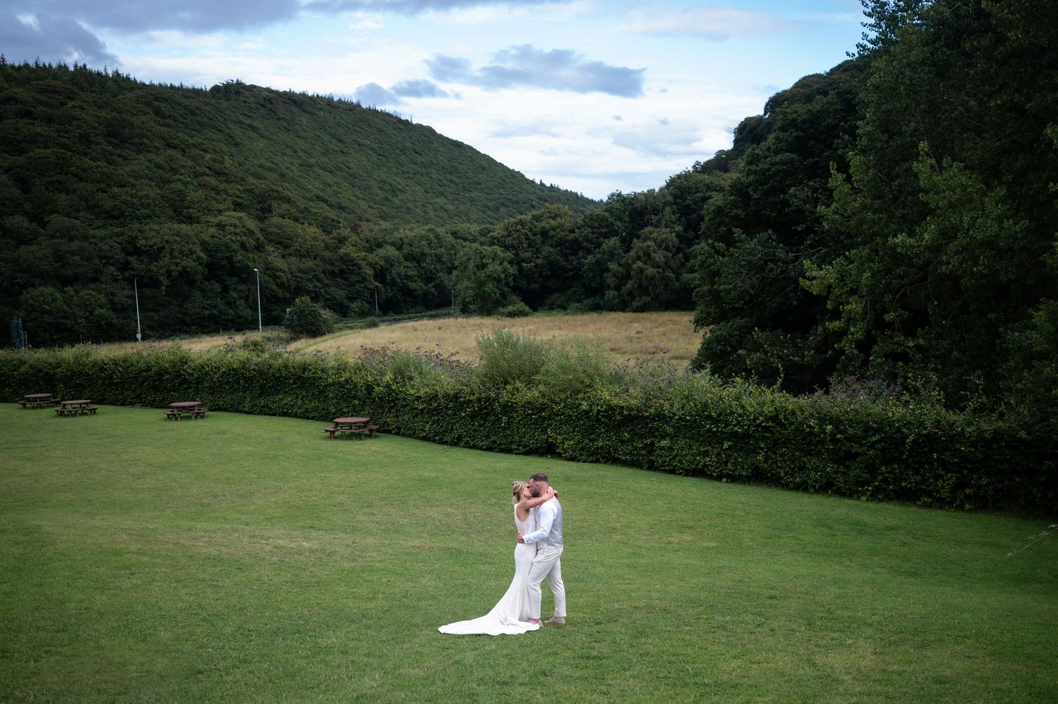 Bride and groom kissing in vast green field with scenic wooded hillside backdrop.