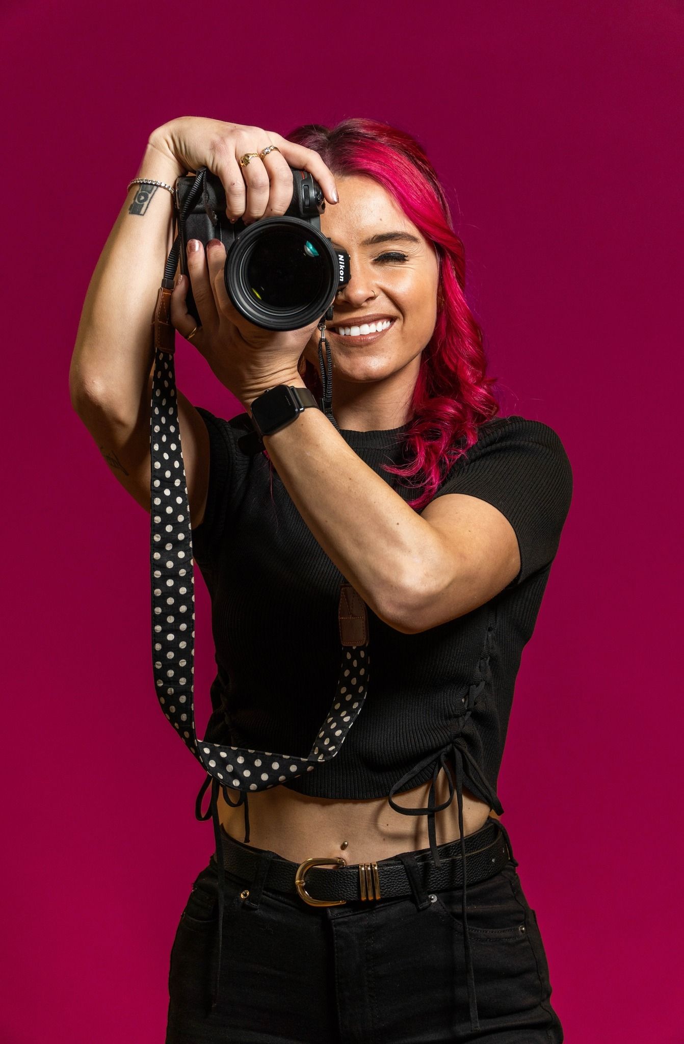 Smiling female photographer with red hair holding DSLR camera on pink background.