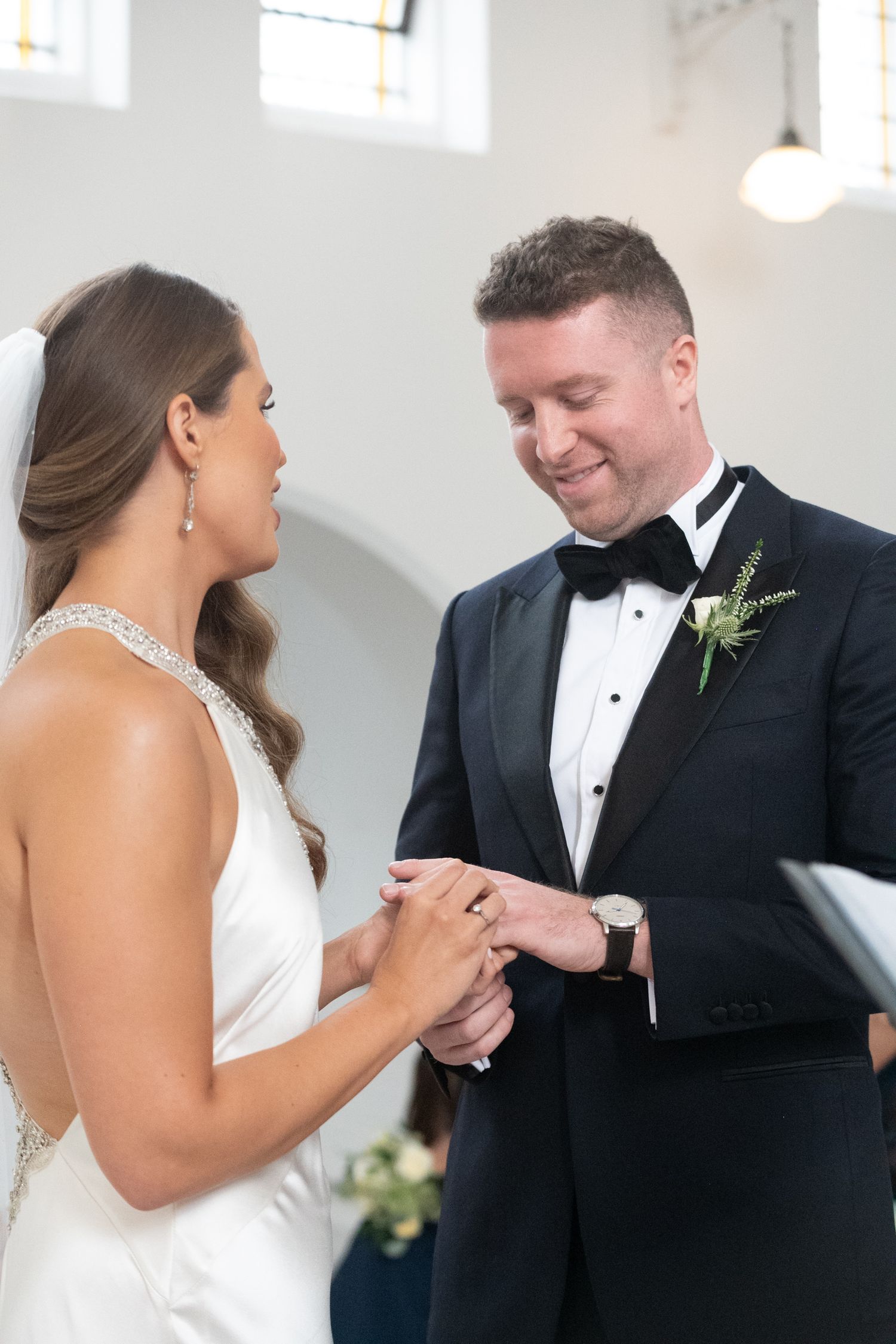 Groom smiling as he places wedding ring on brides finger during church ceremony, both dressed in elegant wedding attire.