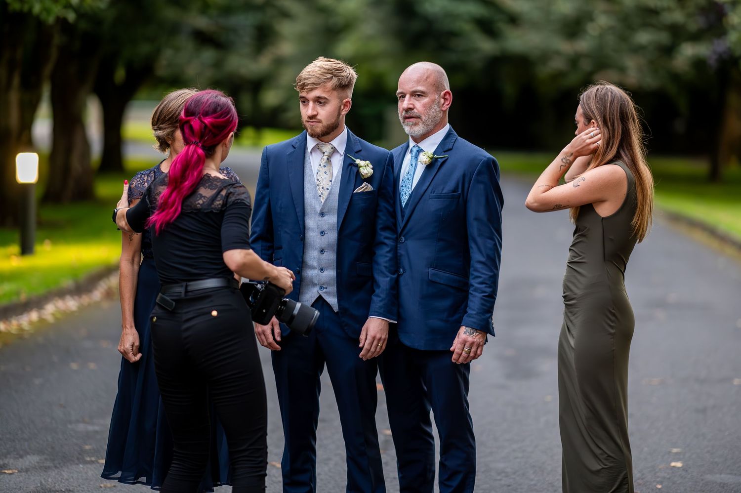 Photographer setting up to photo Two men in blue suits and two women standing on a tree-lined path at a wedding.