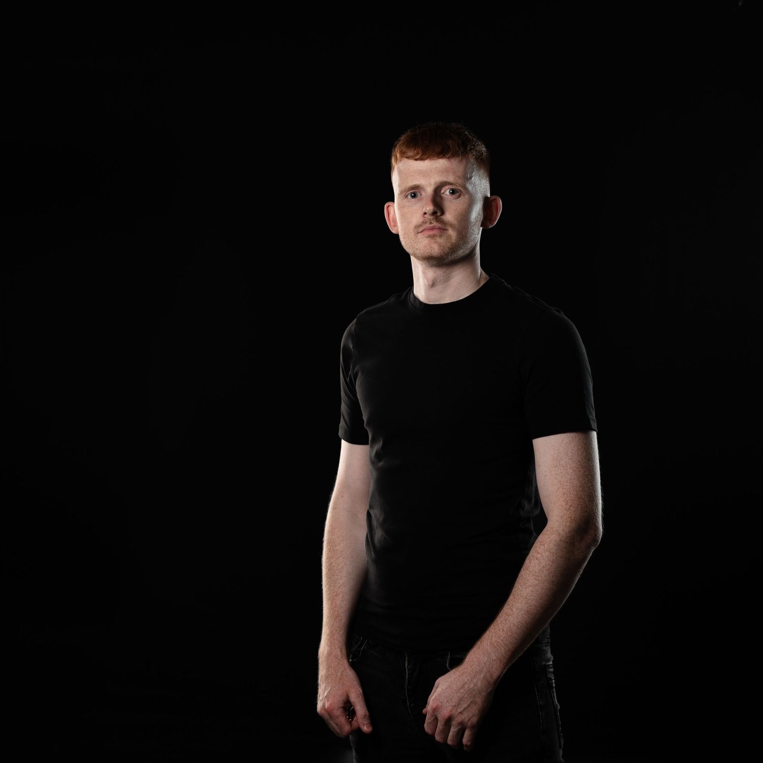 Young man with red hair wearing black t-shirt standing against black background.
