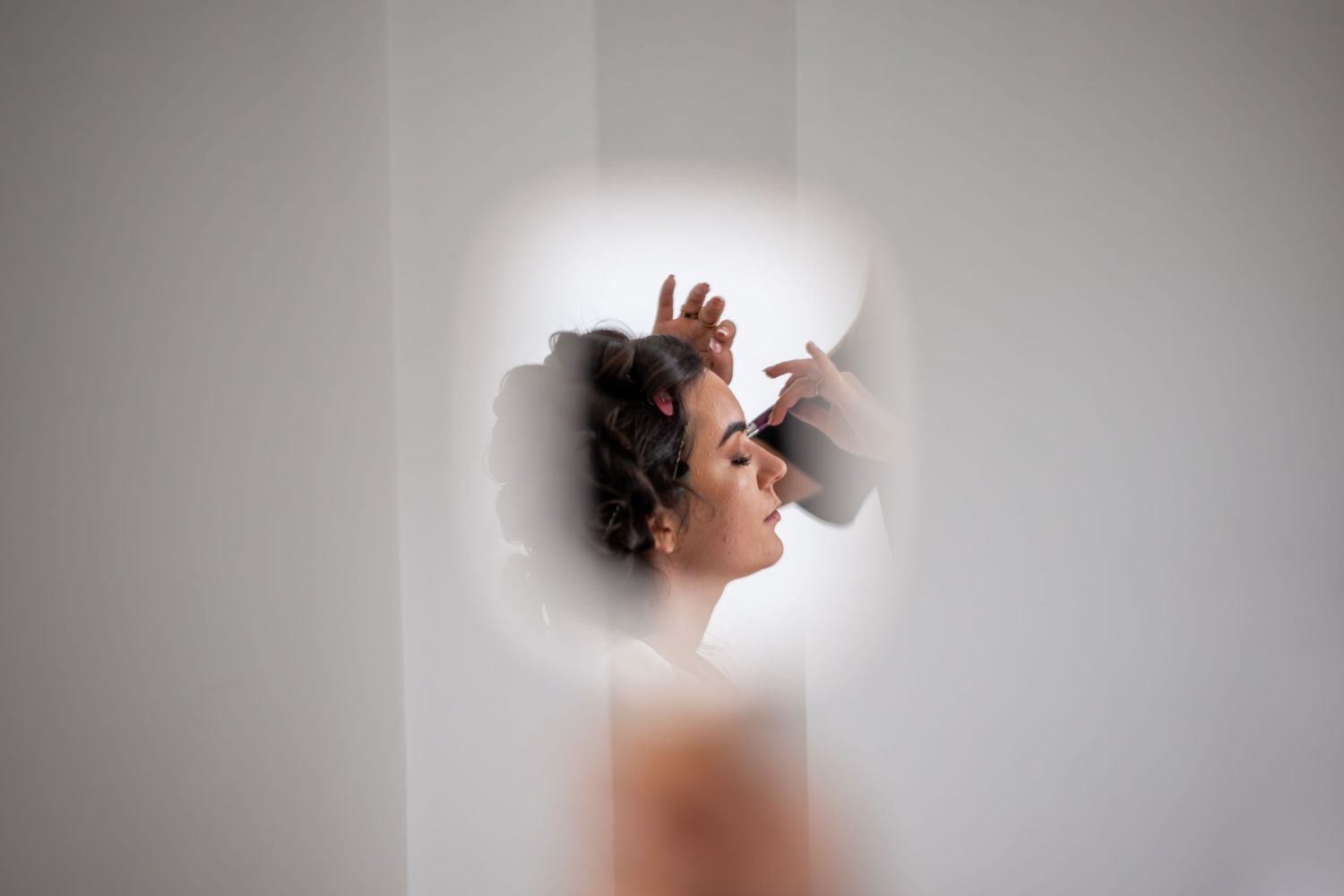 Woman with curly hair viewed through a circular mirror opening, reflecting her profile in soft white light.