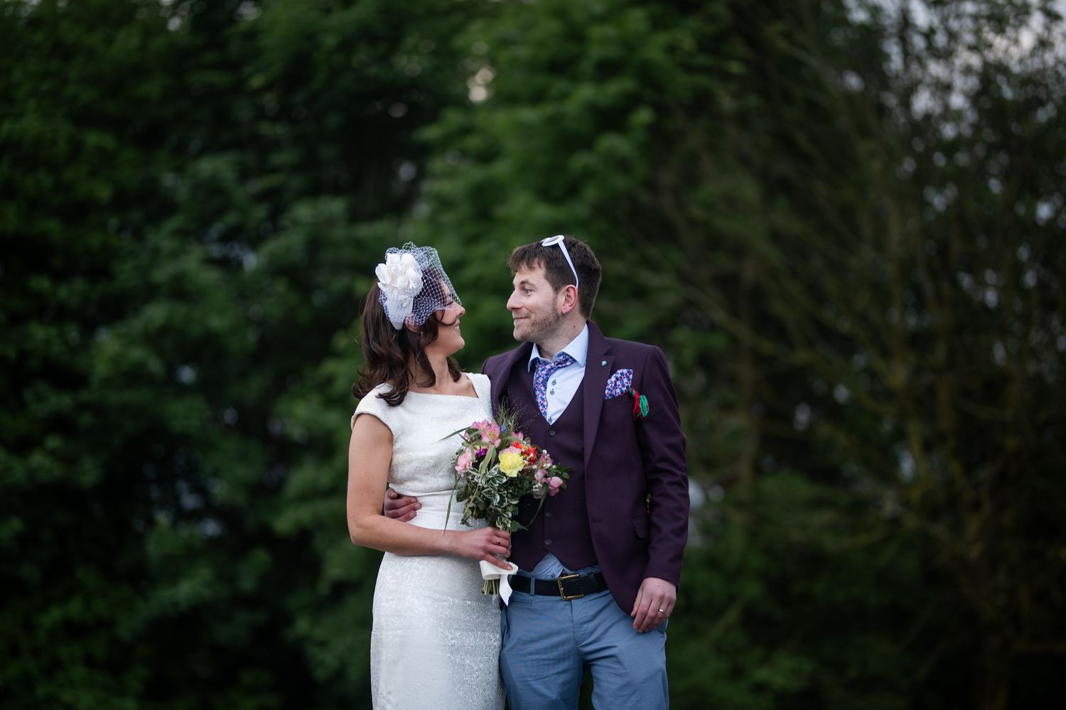 Bride and groom smiling at each other outdoors, bride holding colorful bouquet, groom in purple suit, lush green trees behind.