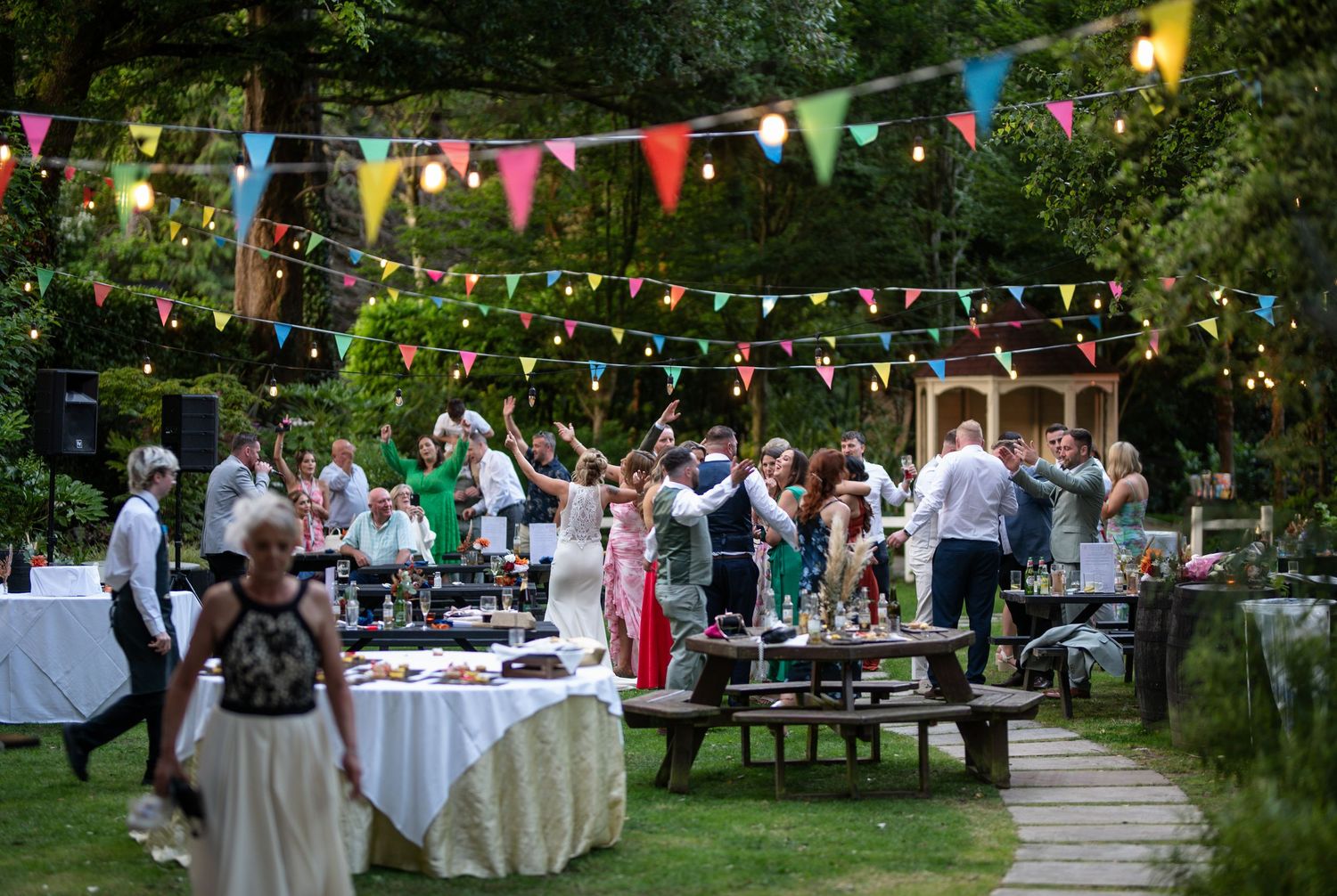 Colorful outdoor wedding reception in garden with string lights and bunting flags.