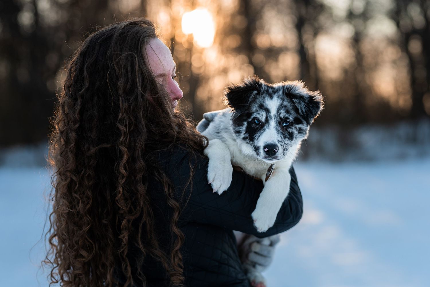 Woman with curly hair holds a black and white puppy outdoors in snowy winter landscape at golden hour sunset.