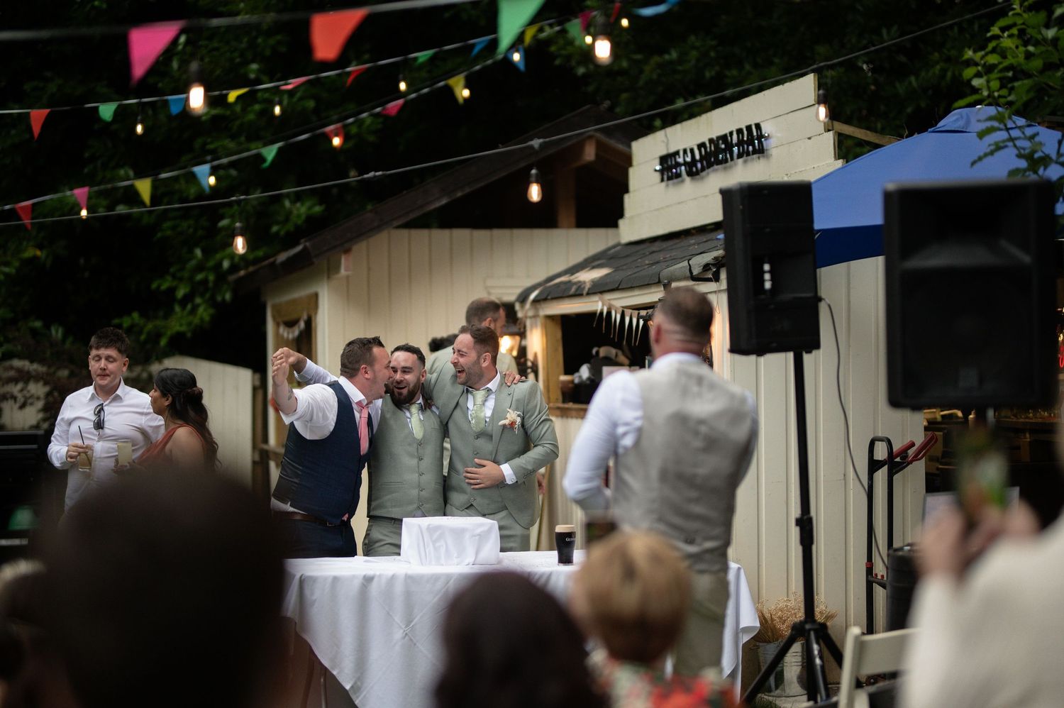 Groom and groomsmen laughing together at outdoor wedding reception with colorful bunting lights.