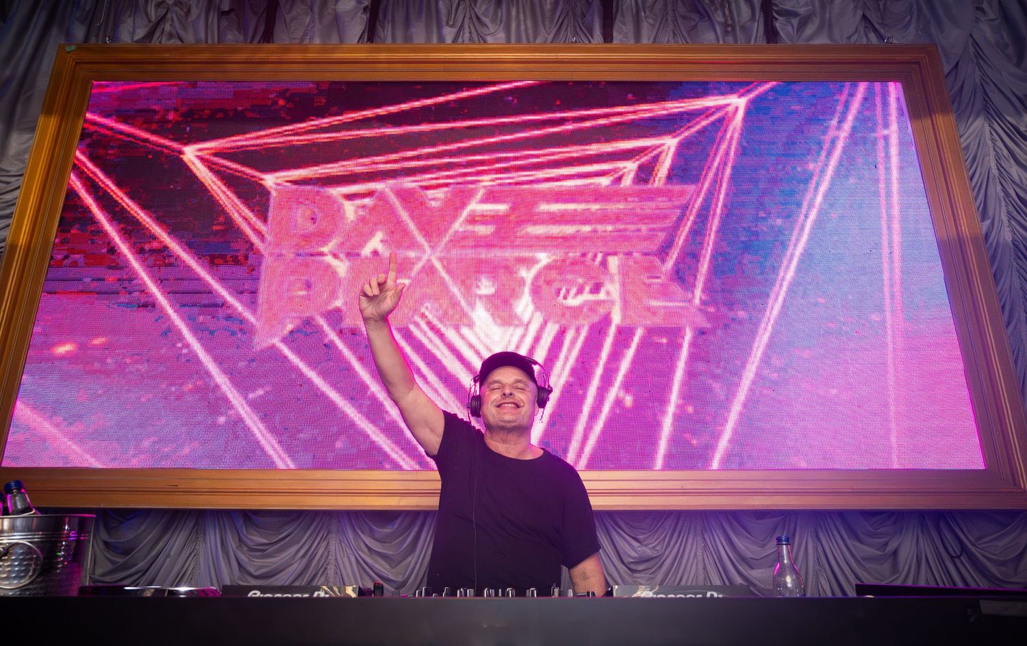 DJ performing on stage with arms raised in front of a vibrant pink laser light display.