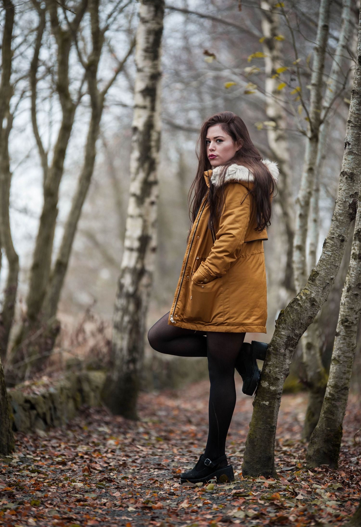 Young woman in mustard coat posing in an autumn woodland forest setting.