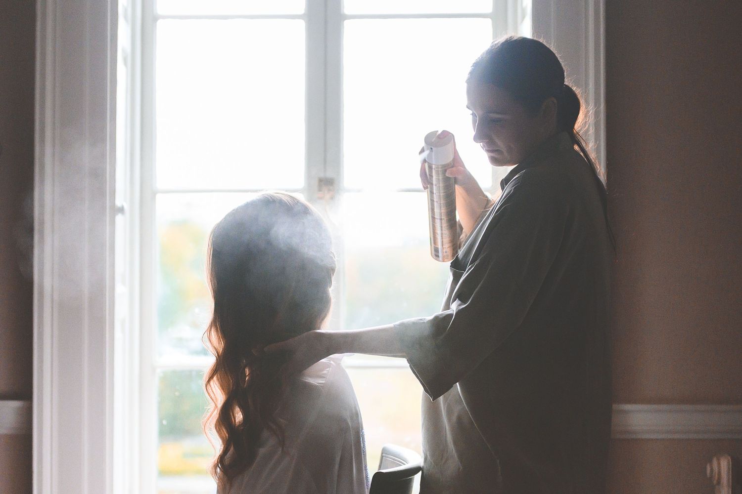 A hairstylist applies hairspray to a bride's hair near a bright window, creating a dreamy backlit silhouette effect.