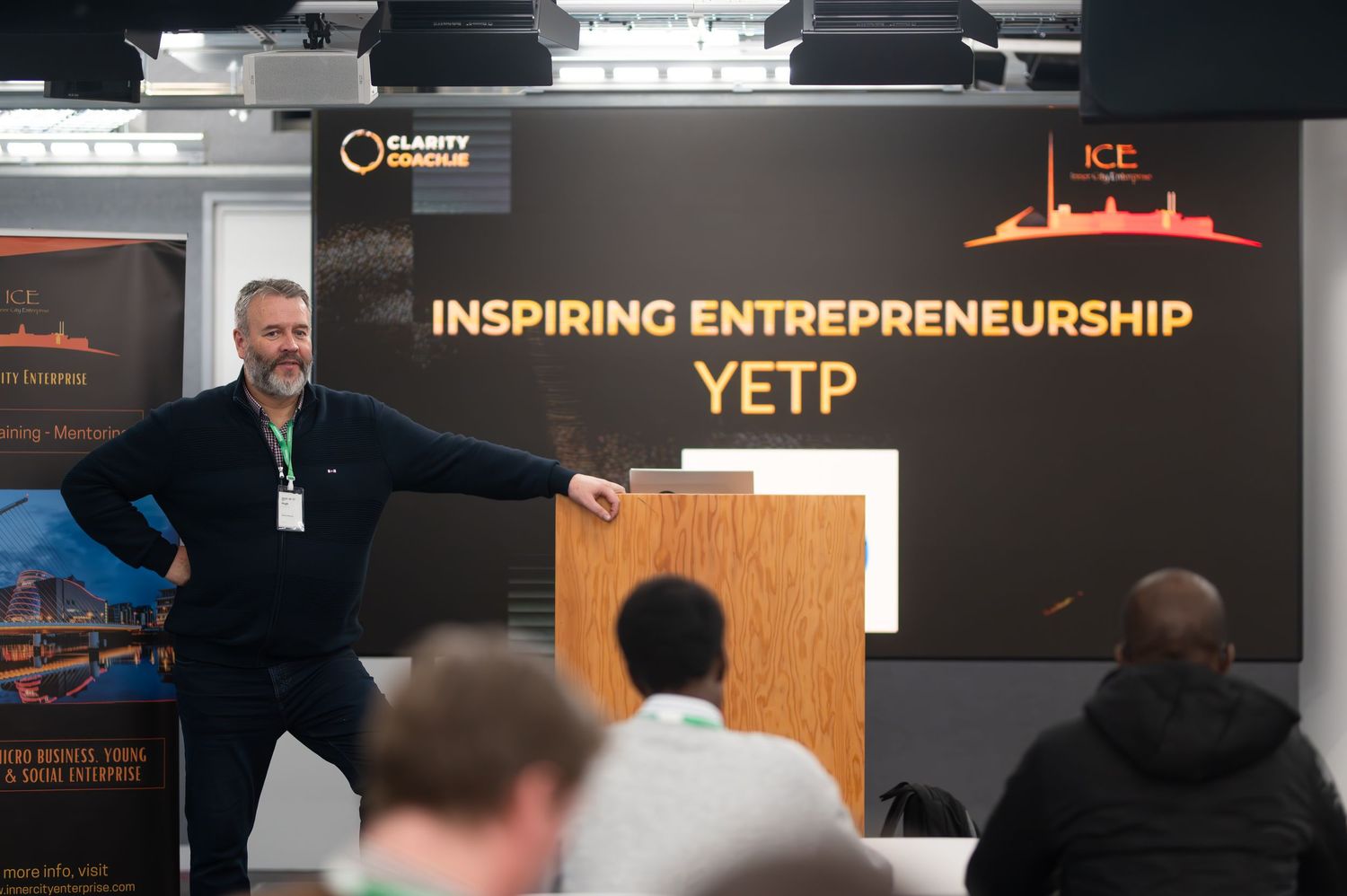 Speaker presenting at YETP Inspiring Entrepreneurship event, gesturing at podium before seated audience.