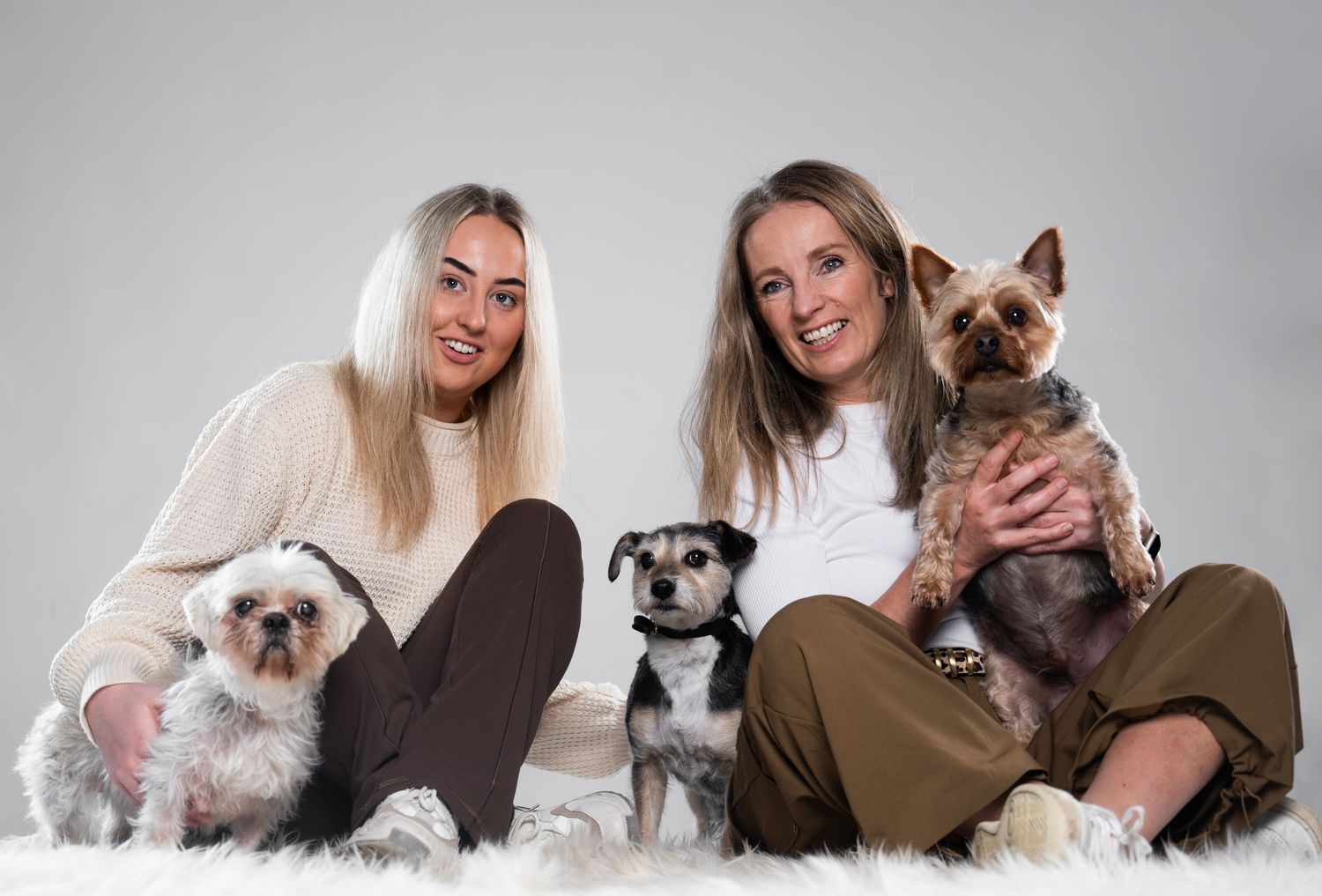 Two blonde women sit cross-legged on a white fur rug, each holding small dogs, with a third dog between them.
