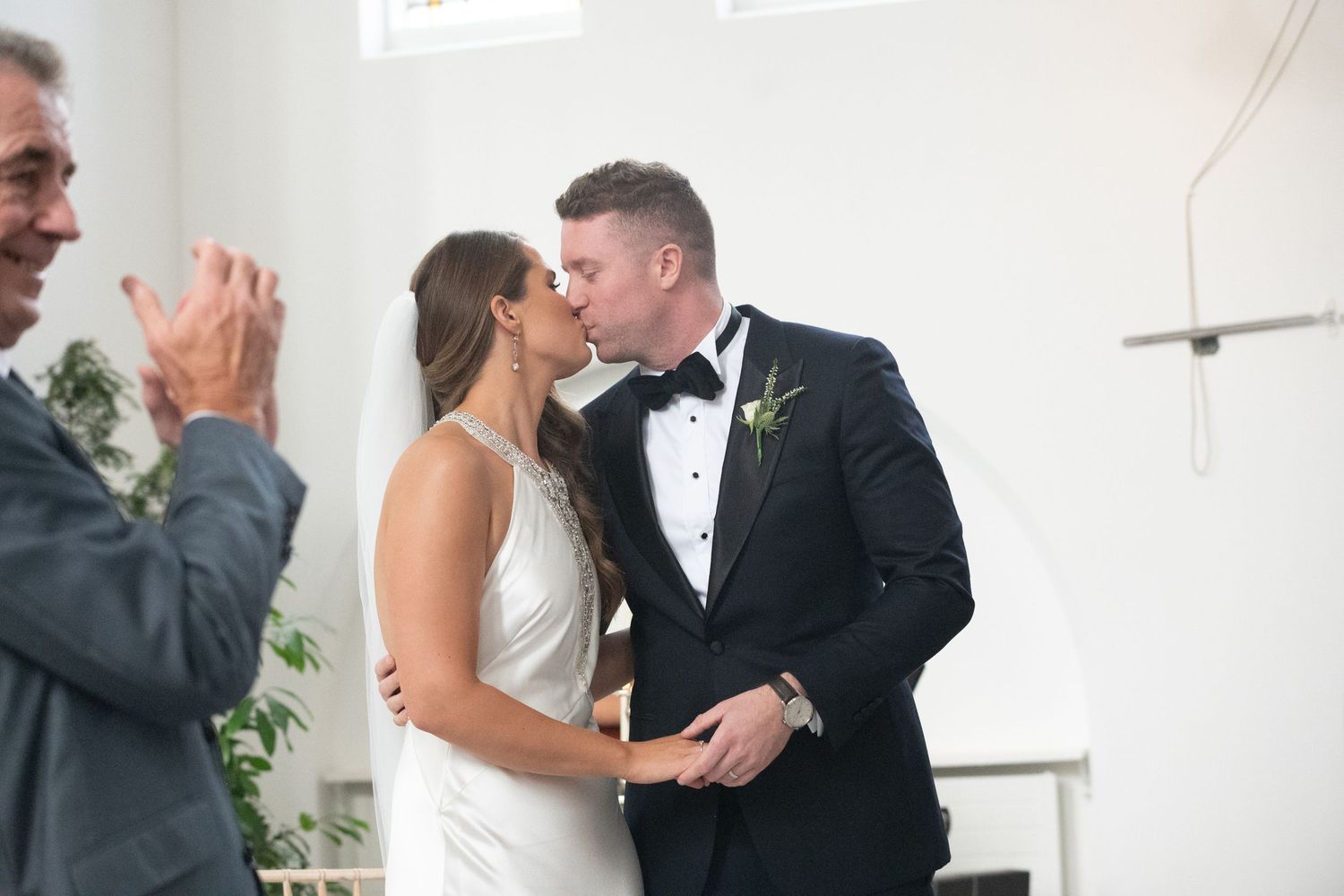 Bride and groom share first kiss at wedding altar as guests applaud in elegant white venue.