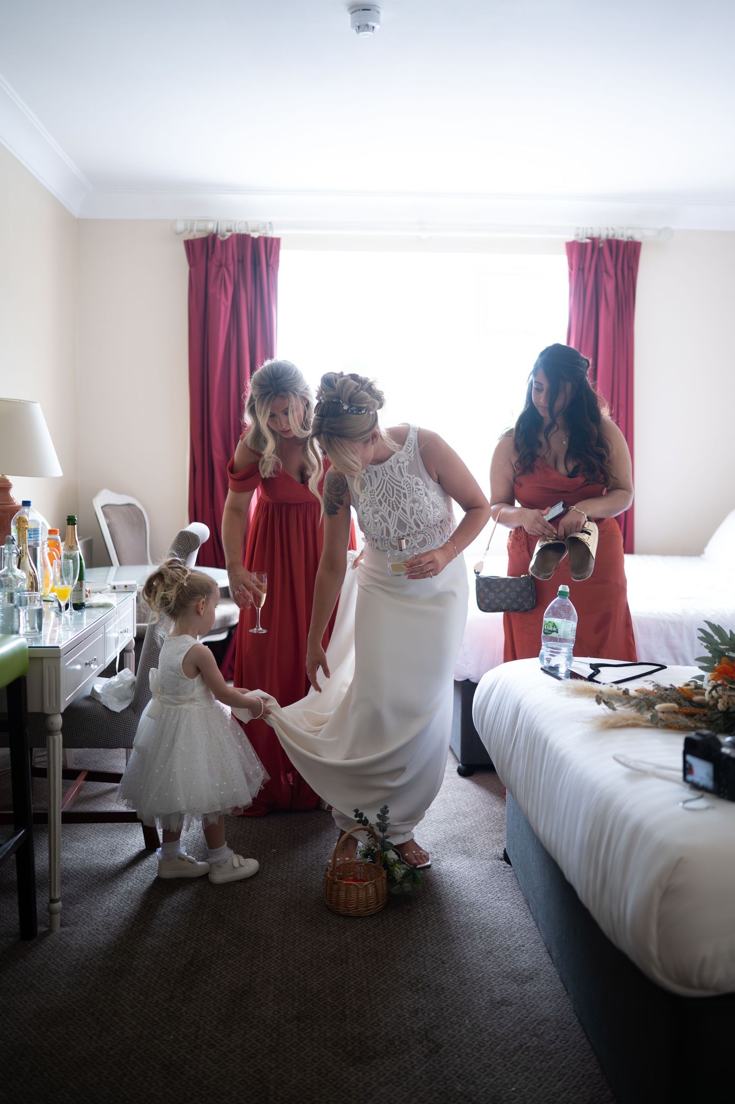 Bride and bridesmaids in red dresses prepare in a hotel room with a flower girl holding the wedding gown.