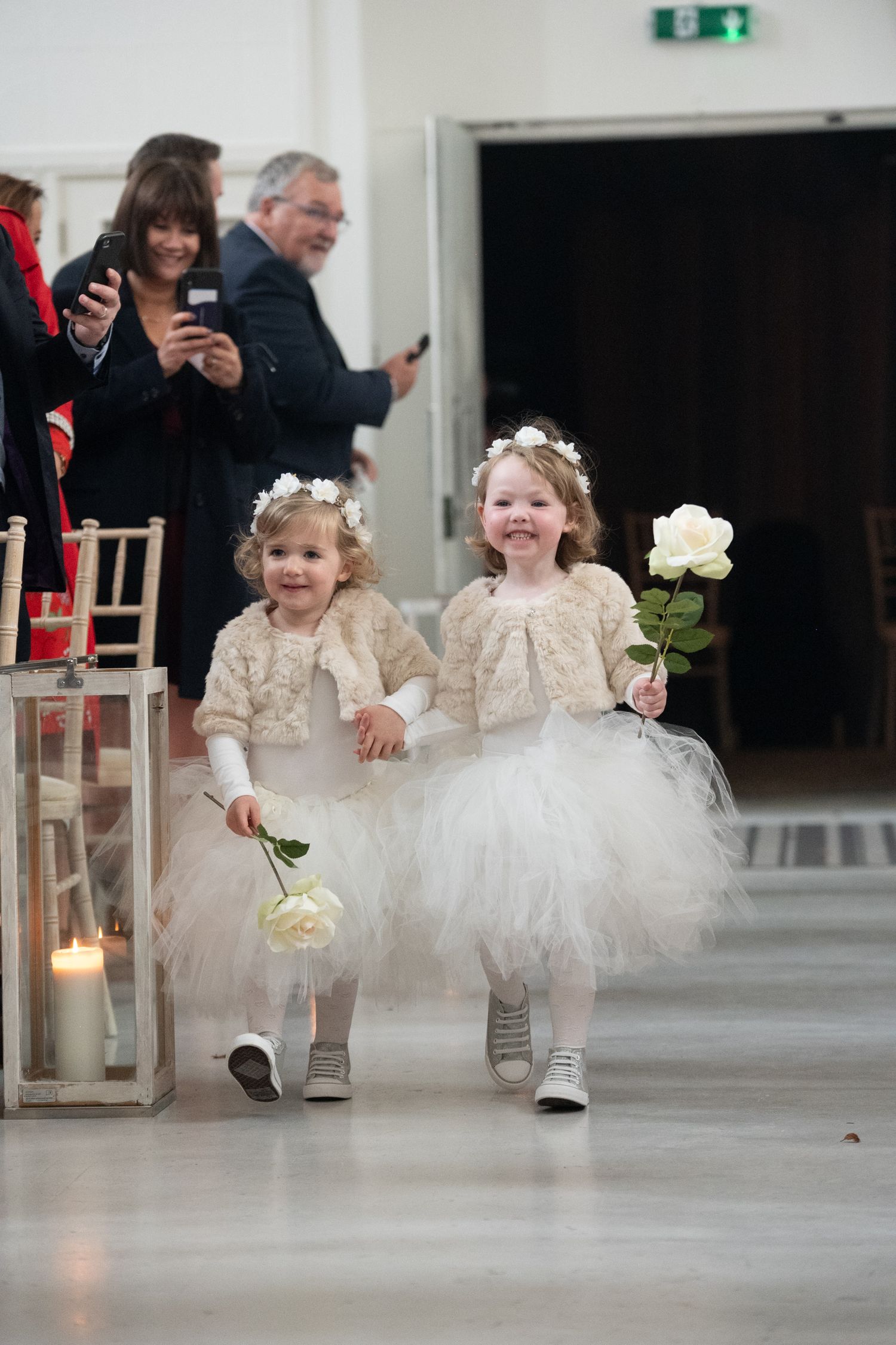 Two adorable flower girls in white tutu dresses and floral crowns walk down the aisle holding white roses at a wedding.