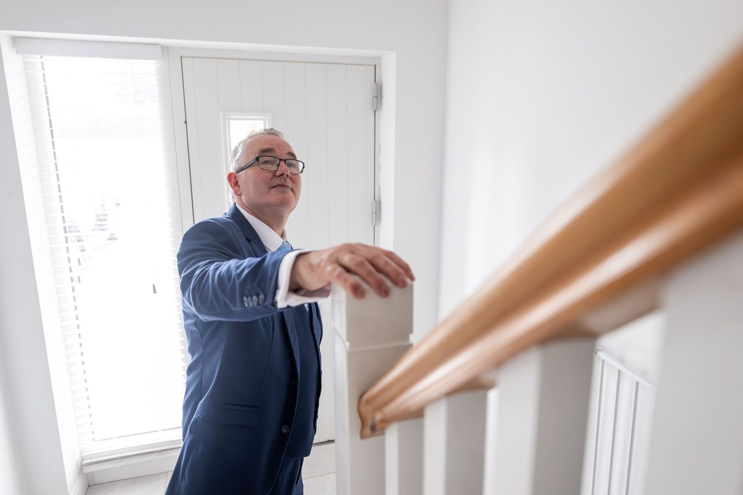 Man in blue suit leaning on wooden stair railing, looking up in a bright modern home interior.