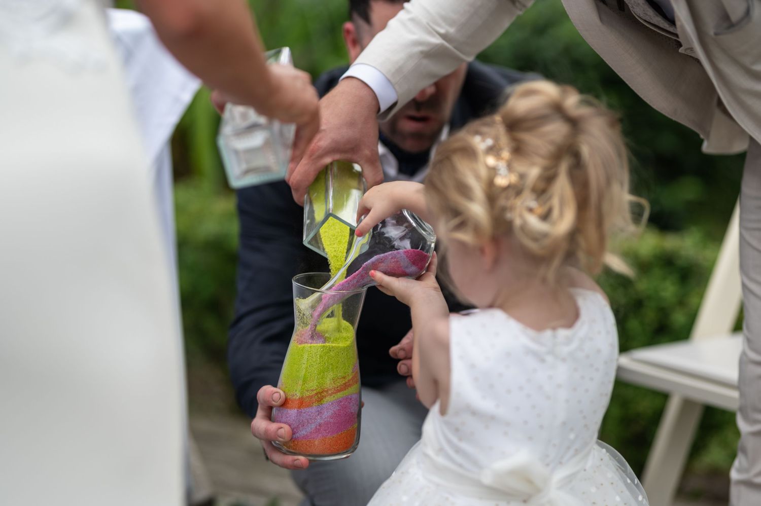 Young flower girl participates in colorful sand unity ceremony during outdoor wedding celebration.