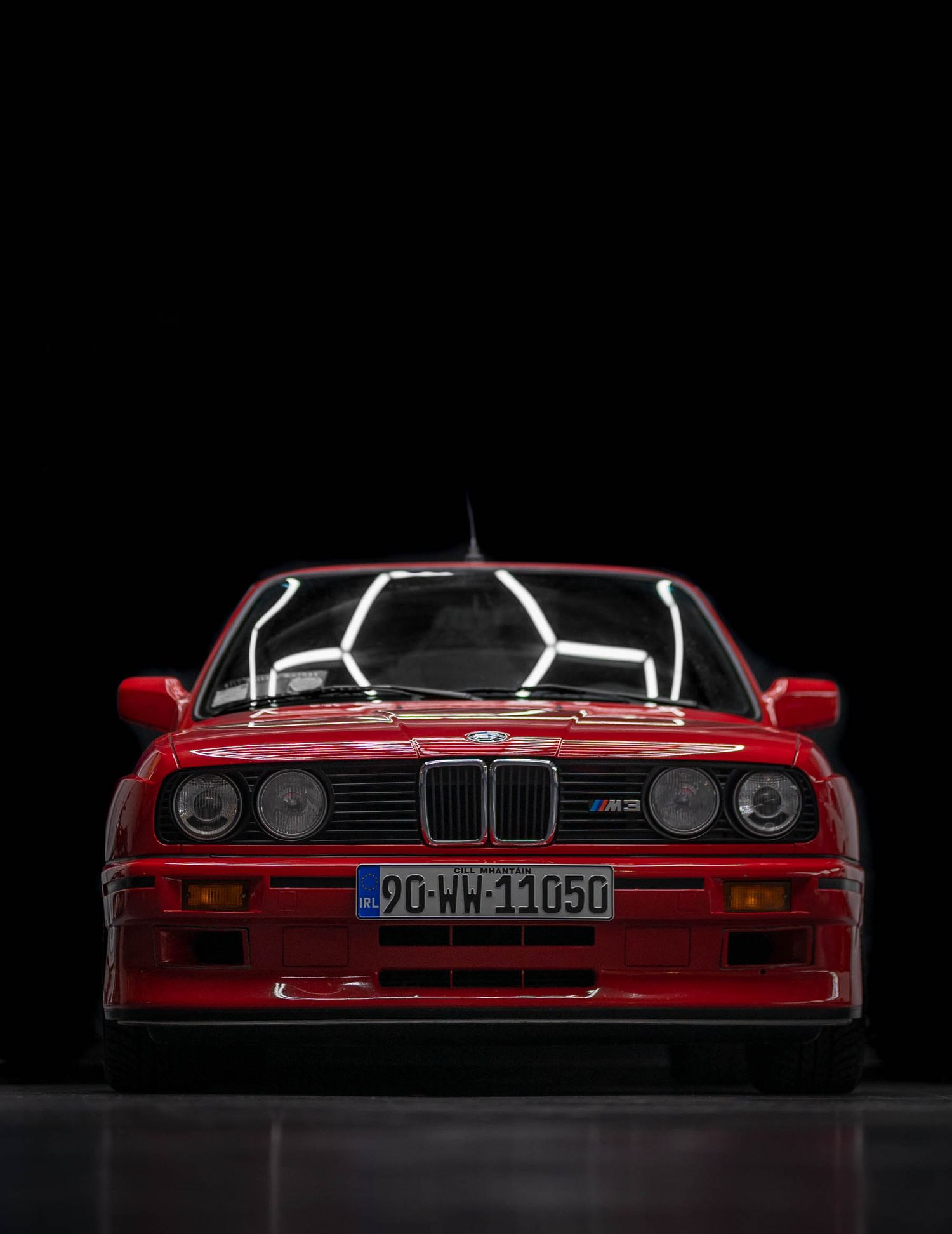 Classic red BMW M3 E30 photographed front-on against black studio background.