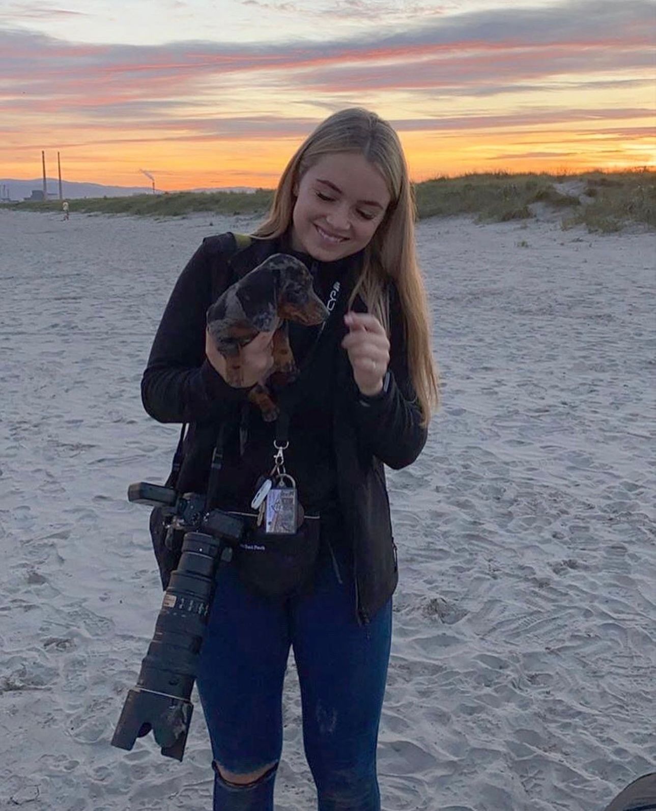 Smiling young woman holding dachshund puppy on beach at sunset with professional camera.
