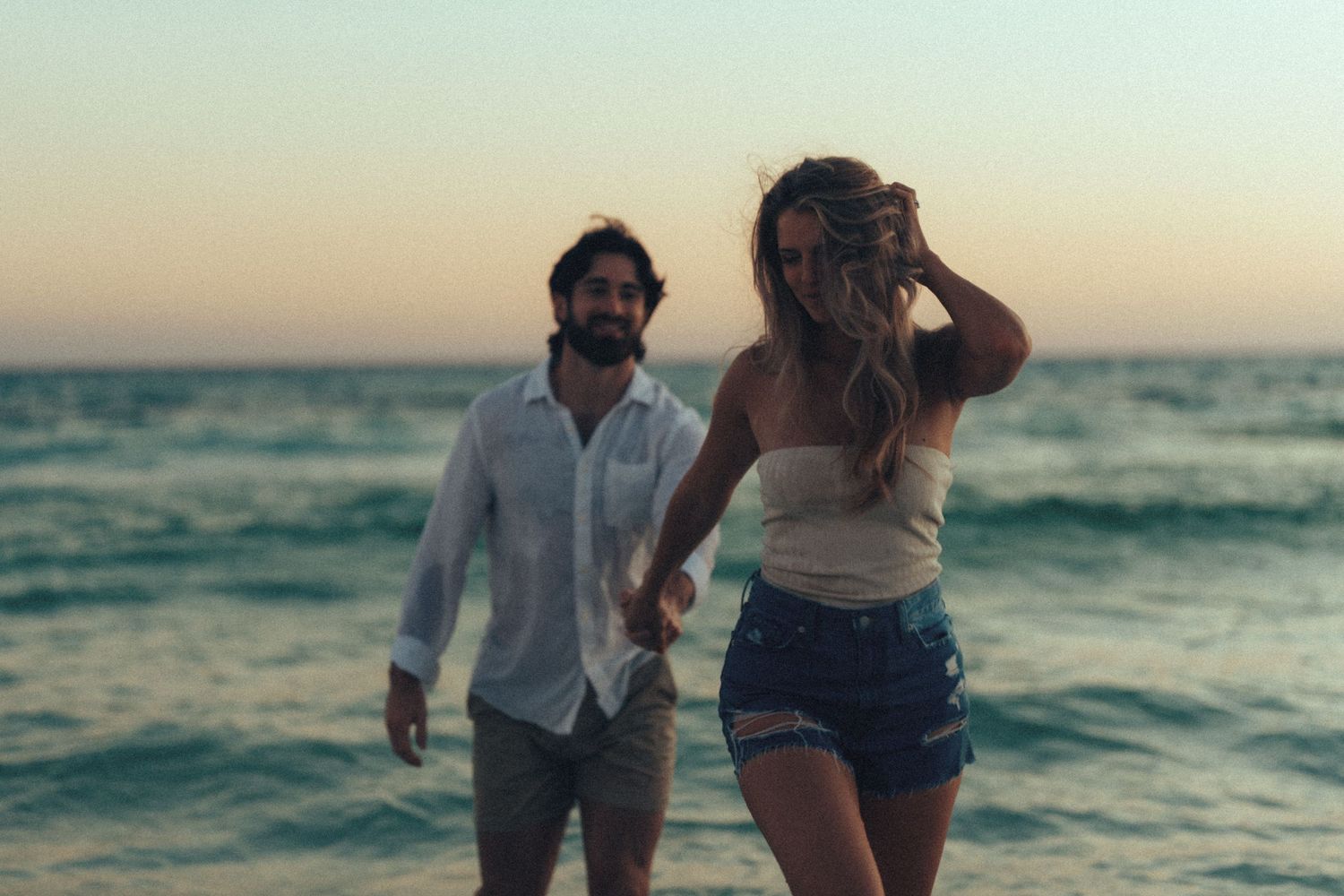 A couple walks along the beach at sunset with ocean waves in the background.