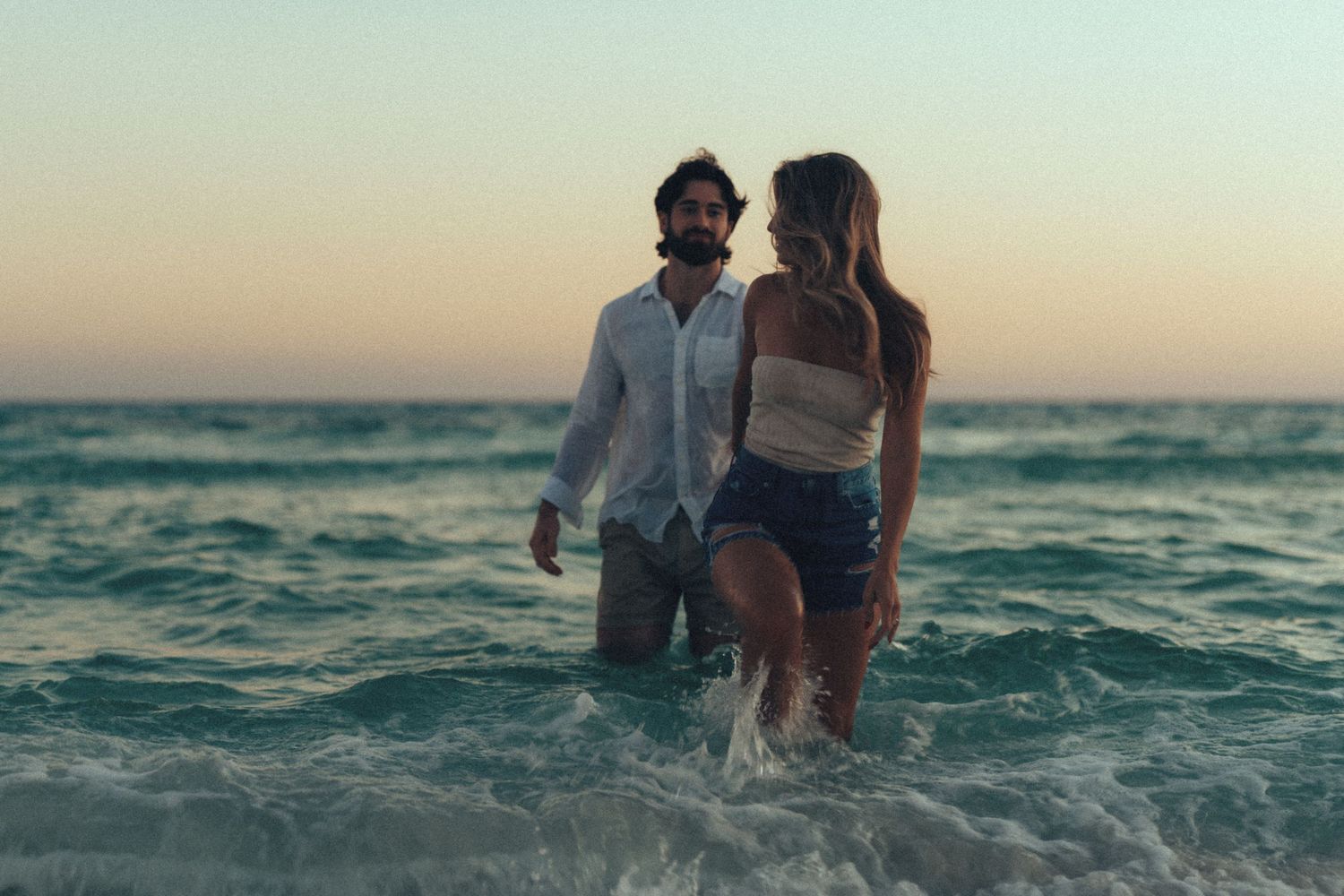 A couple walks together in shallow ocean waves at sunset creating a romantic silhouette against the horizon.