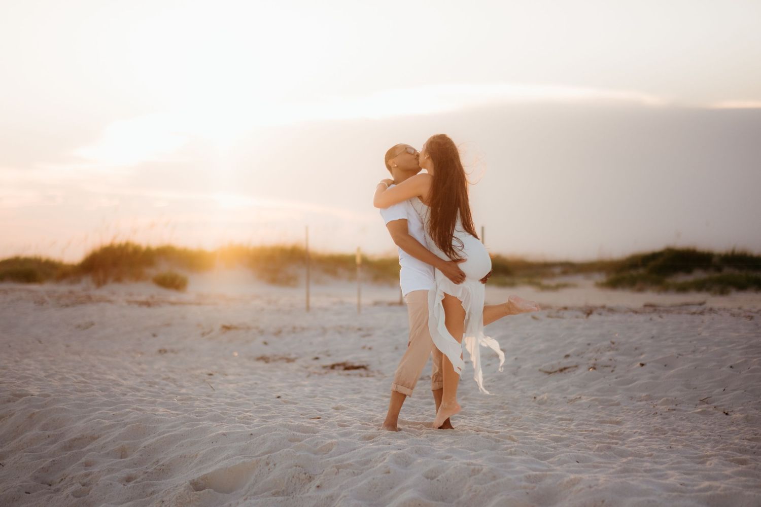 A silhouetted couple shares an intimate moment on sandy dunes at sunset.