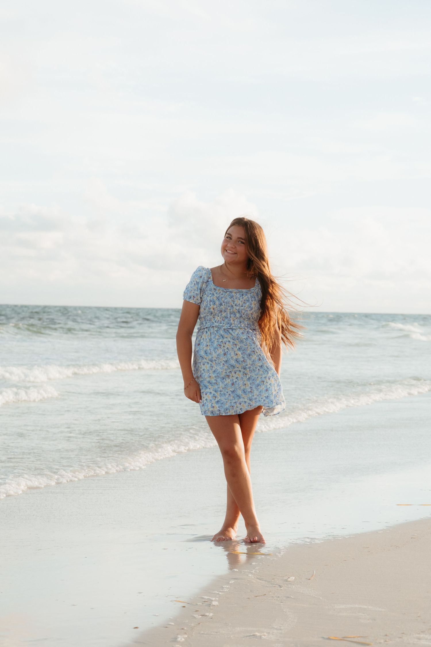 Person in a blue floral dress walking along a tranquil beach shoreline.