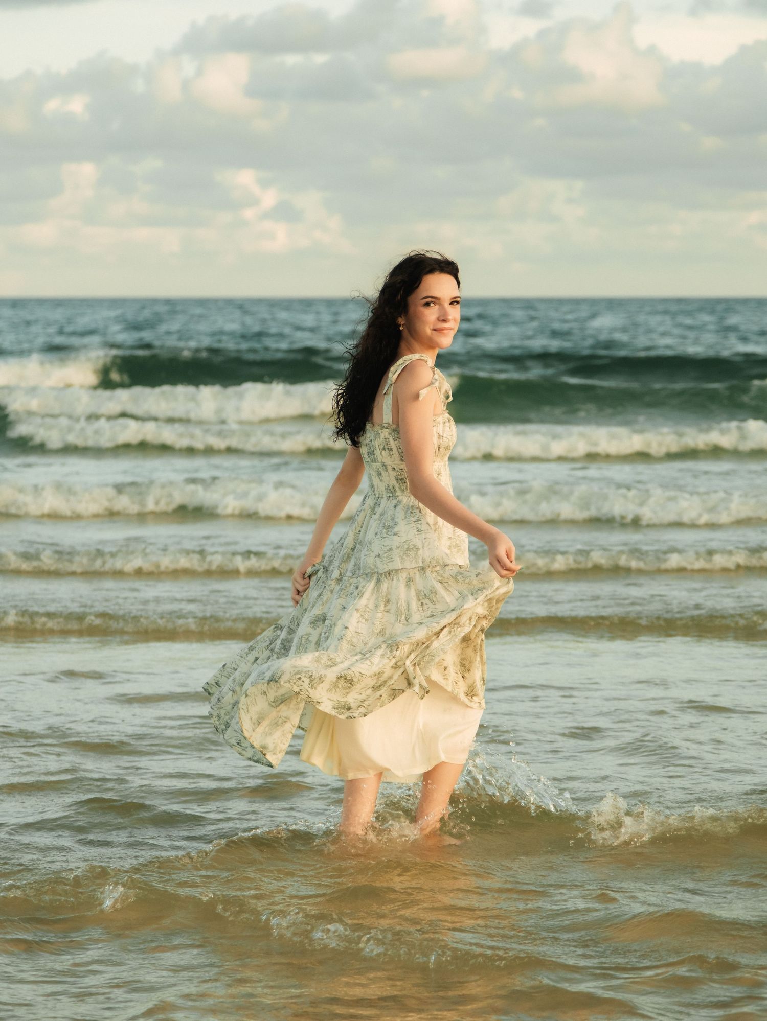Woman in flowing dress twirling and splashing in ocean waves at sunset.