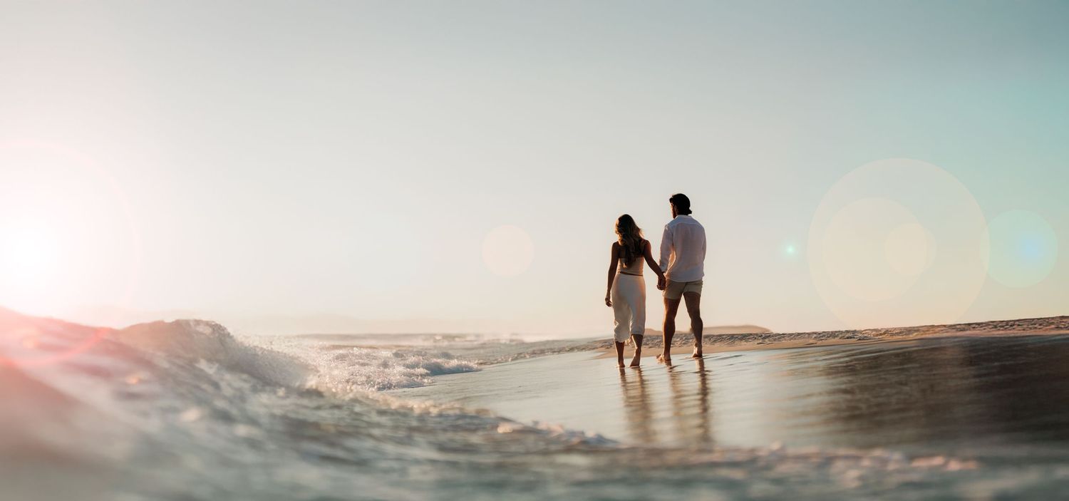 Two silhouettes stand together on a beach during sunset with water washing over their feet.