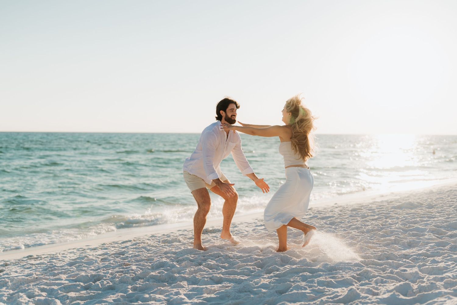 A couple playfully splashes in the ocean waves at sunset on a white sandy beach.