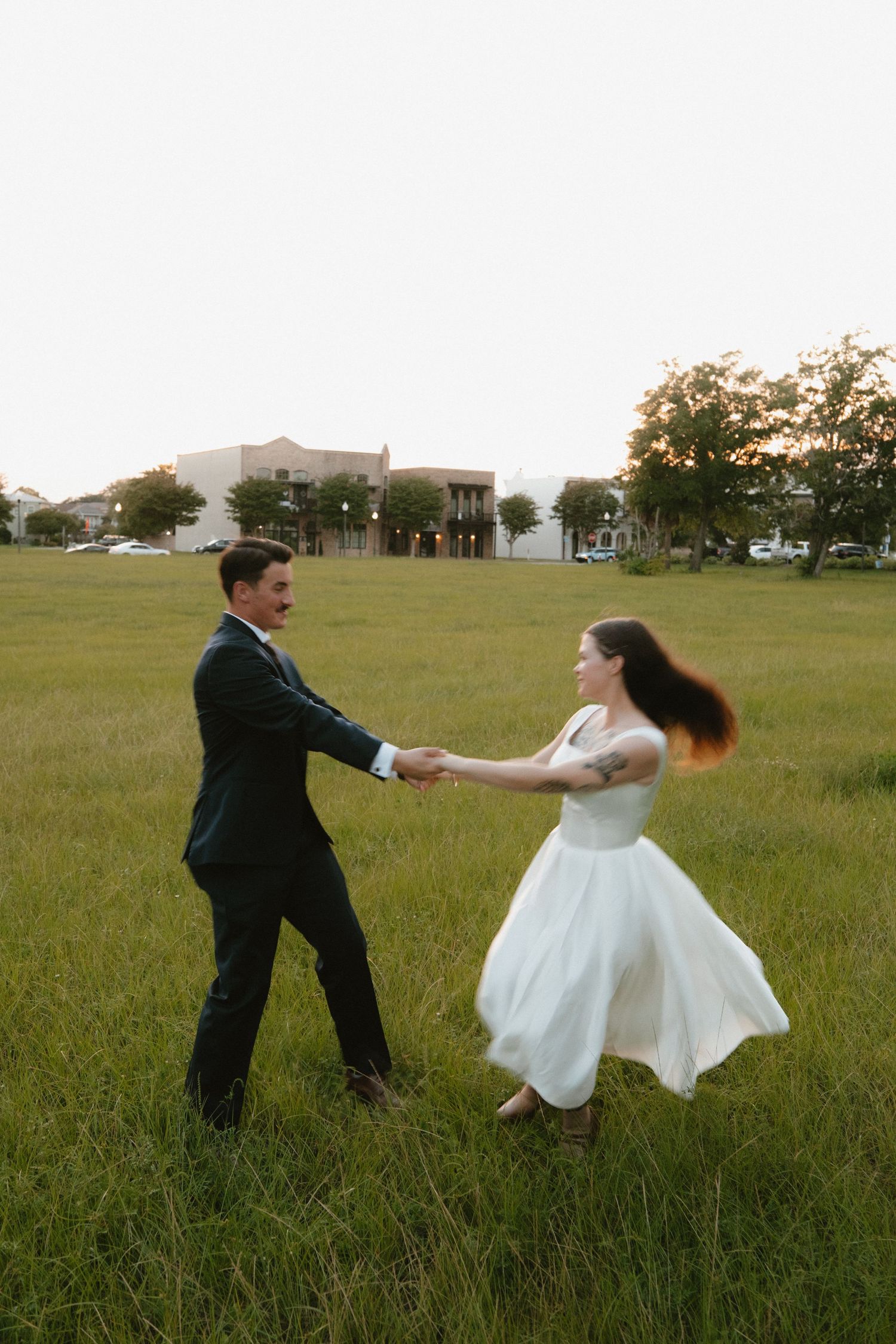 A couple dances playfully in a grassy field wearing formal wedding attire.