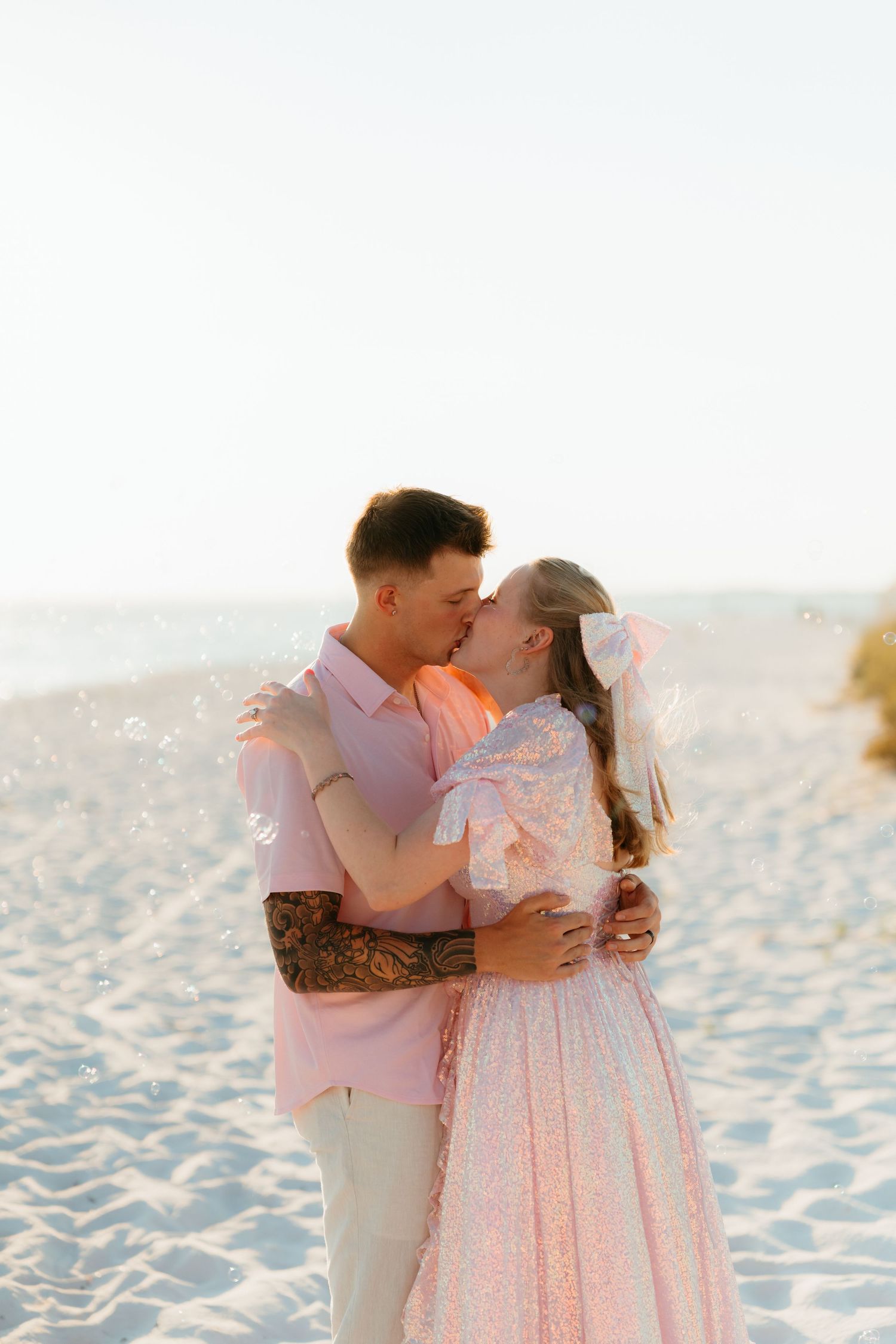 A romantic couple shares a tender kiss on a white sandy beach at sunset while wearing coordinating pink and white outfits.