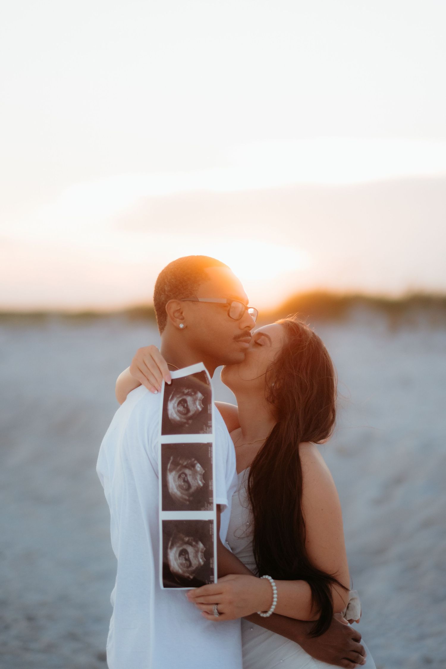A couple sharing a tender moment on the beach while holding ultrasound photos.